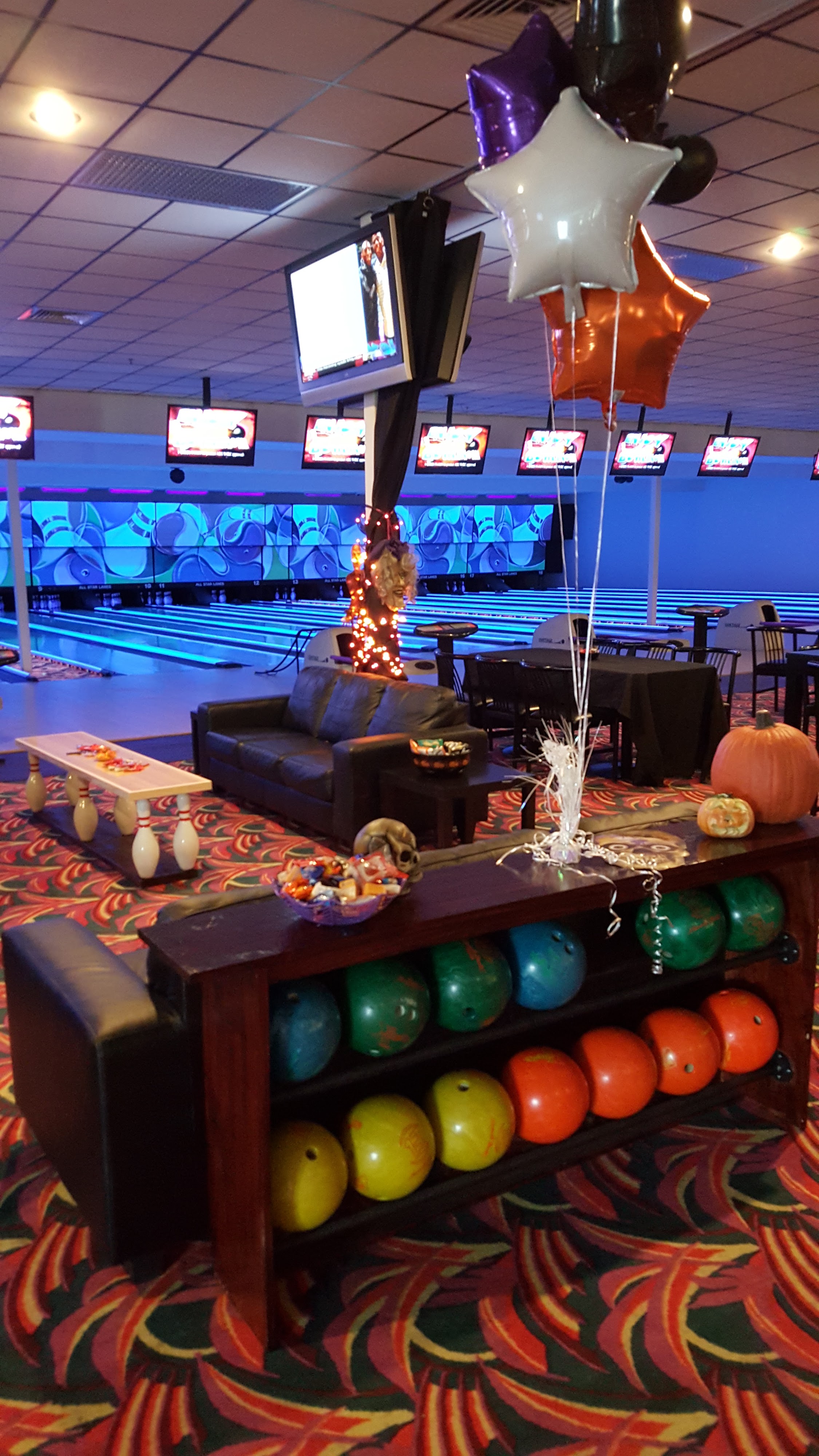A bowling alley lounge area decorated for Halloween with pumpkins, balloons, and colorful bowling balls on a rack.