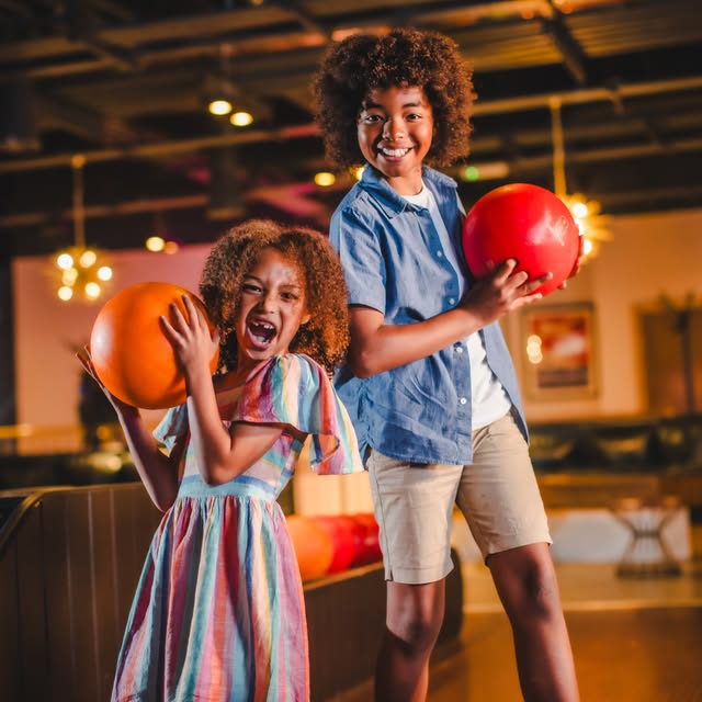 Two children, a boy and a girl, smiling and holding bowling balls in a bowling alley.