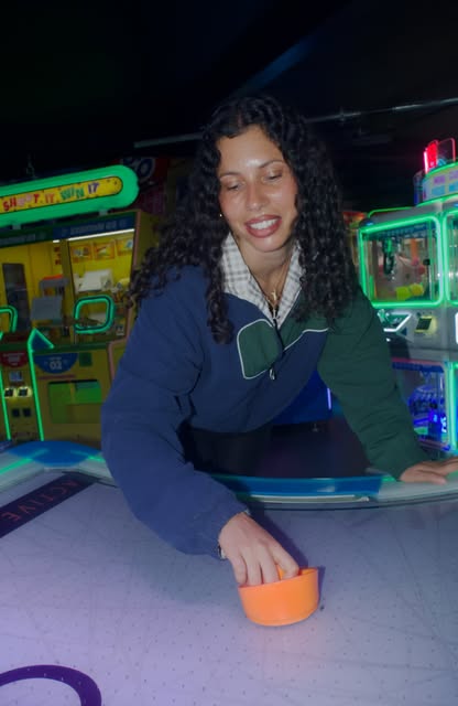 A smiling woman with curly hair plays air hockey in a dimly lit arcade surrounded by glowing game machines.