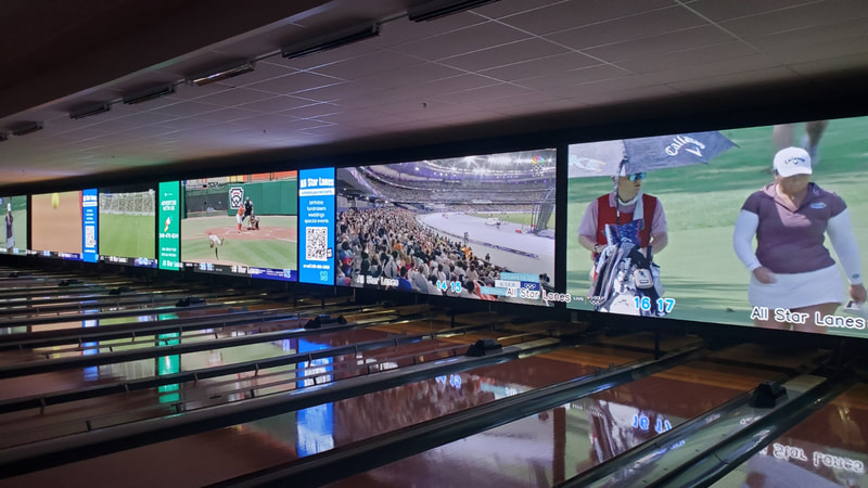 A long row of bowling lanes with massive overhead digital screens showing sports broadcasts.