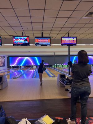 A person films a friend bowling on a lane with purple neon lighting and overhead scoring monitors.