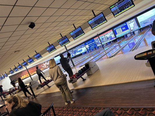 A busy bowling center with people standing at the lanes and many overhead scoring monitors.