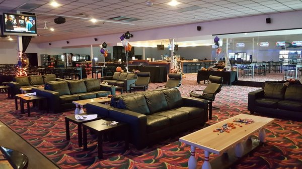 A spacious bowling alley lounge with black leather couches, bowling pin coffee tables, and an adjacent bar area.