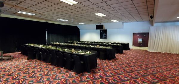 Large event room with long banquet tables set with black linens and green decor on patterned carpet.