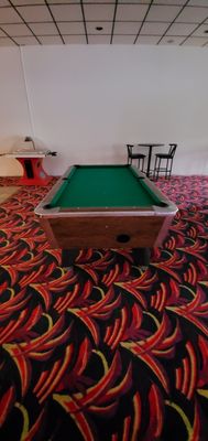 A green felt pool table stands on patterned carpet near a foosball table and high-top seating.
