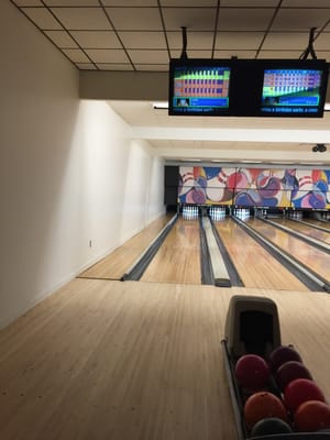 A view of bowling lanes from the approach showing a ball return and overhead scoring monitors.