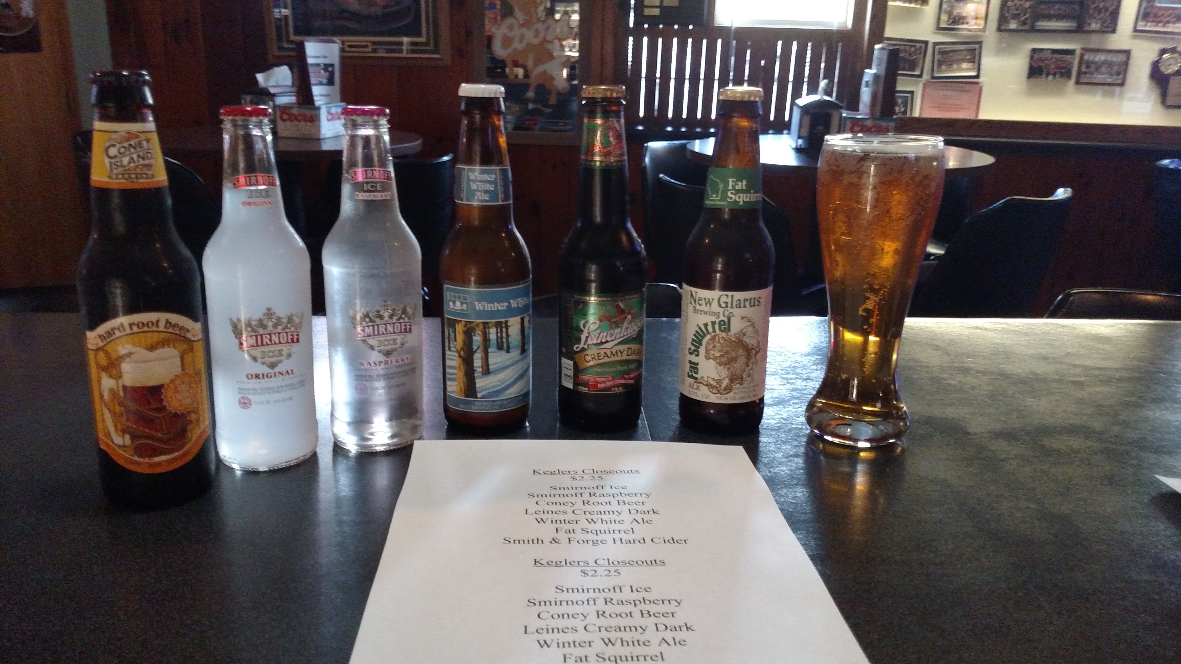 A close-up photo shows six bottles of beer and a glass of beer arranged on a dark table next to a paper menu.