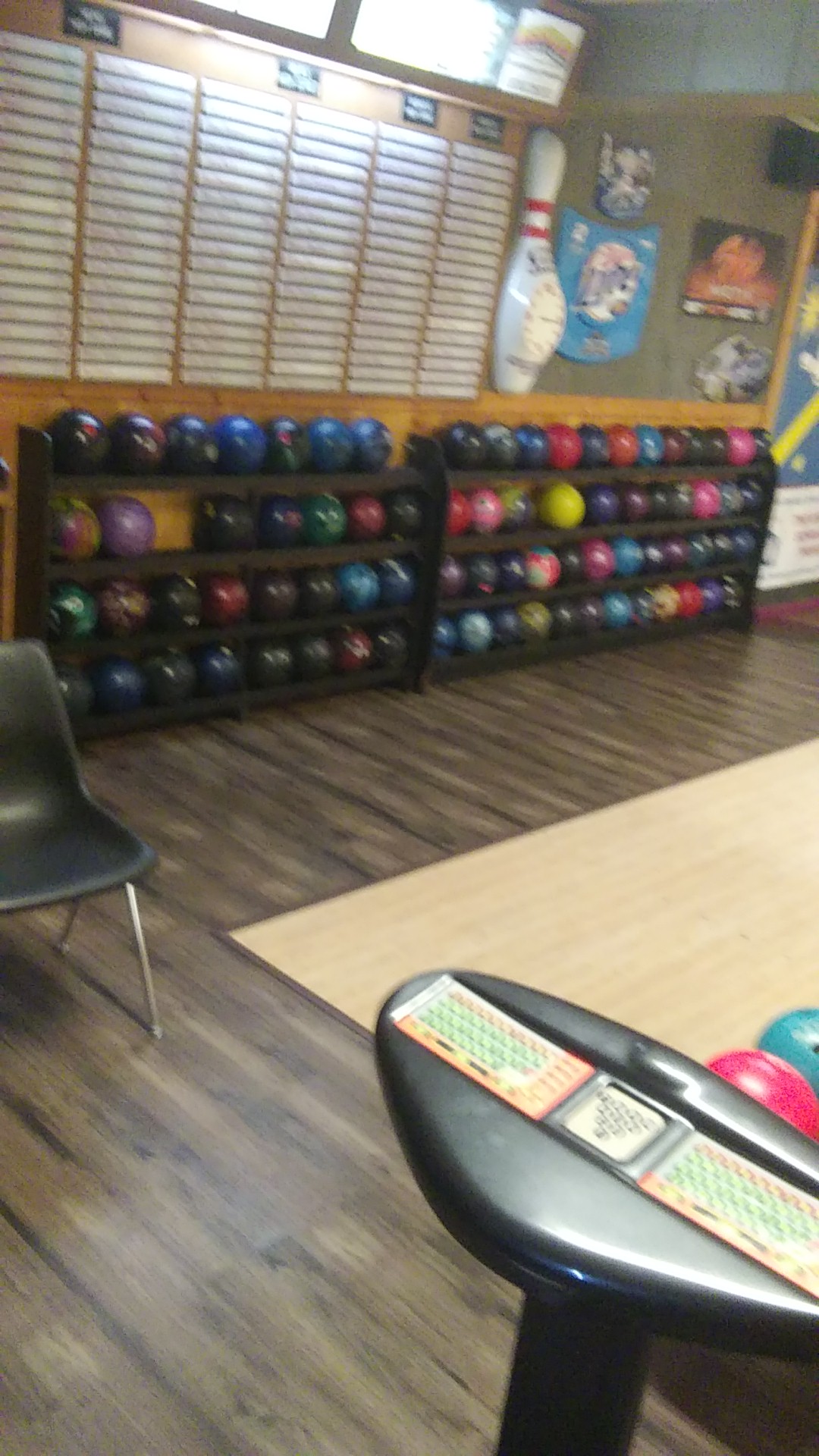 A dimly lit bowling alley showing ball racks filled with colorful bowling balls, a black chair, and a ball return.