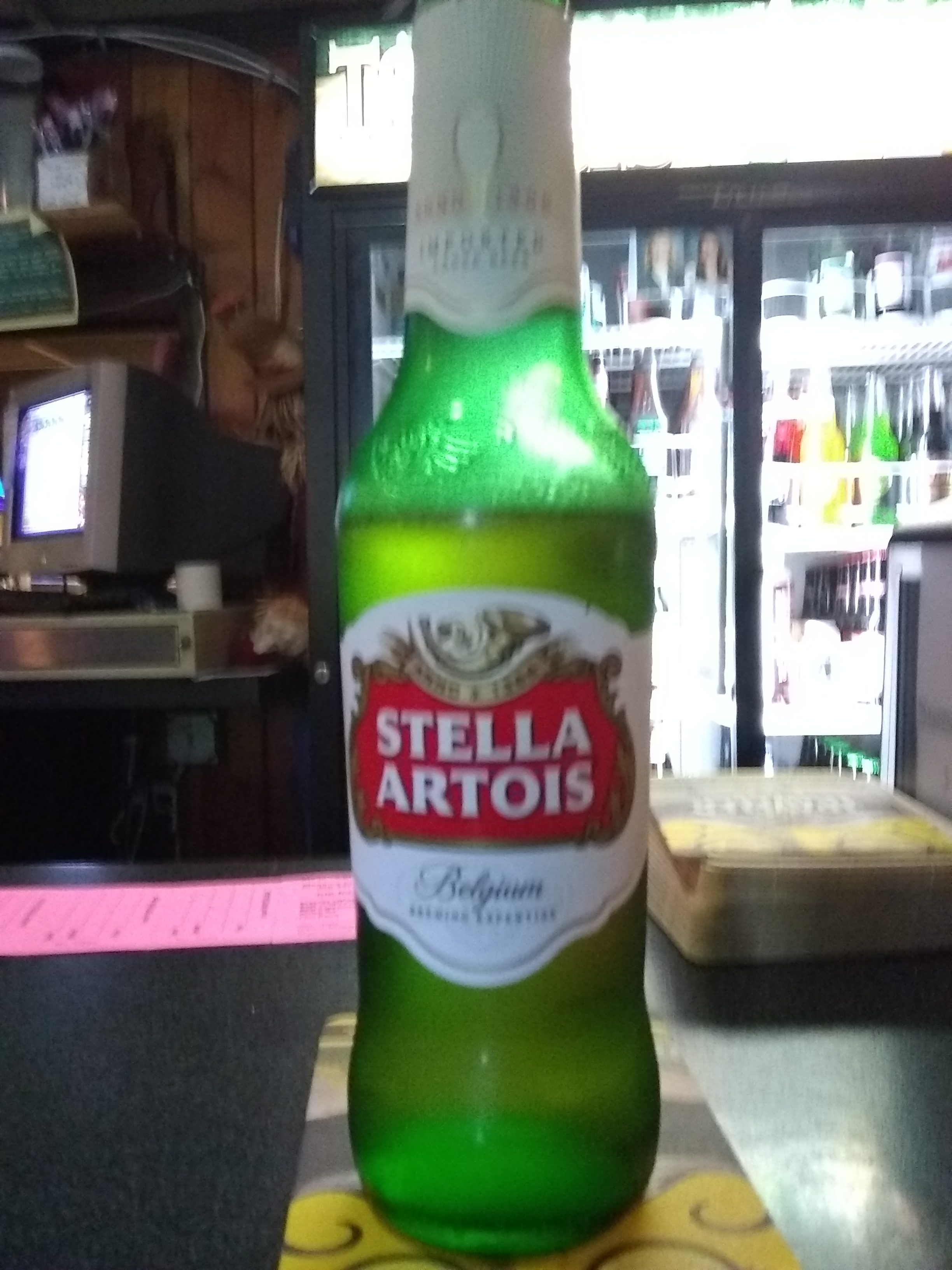 A Stella Artois beer bottle on a yellow coaster, with a blurred background of a bar area and drink cooler.