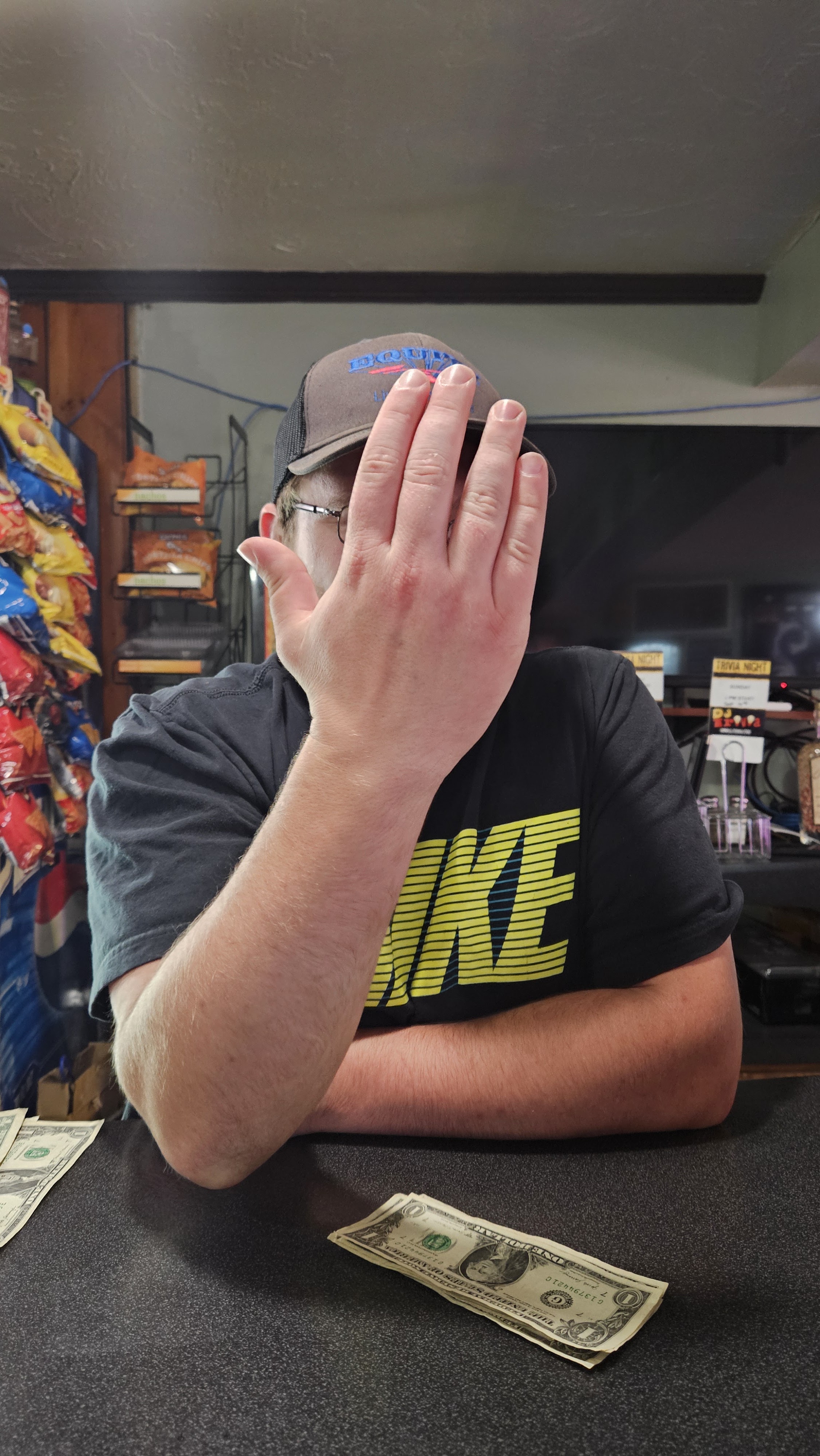 A man in a baseball cap and glasses sits at a dark counter, holding his hand over his face, with snack bags and dollar bills visible.