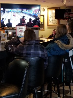 People watch a bowling game on a large television screen above a bar, with framed art on the wall.