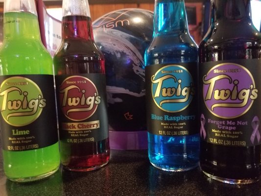 Four colorful bottles of Twig's soda are on a dark counter with a bowling ball in the blurred background.