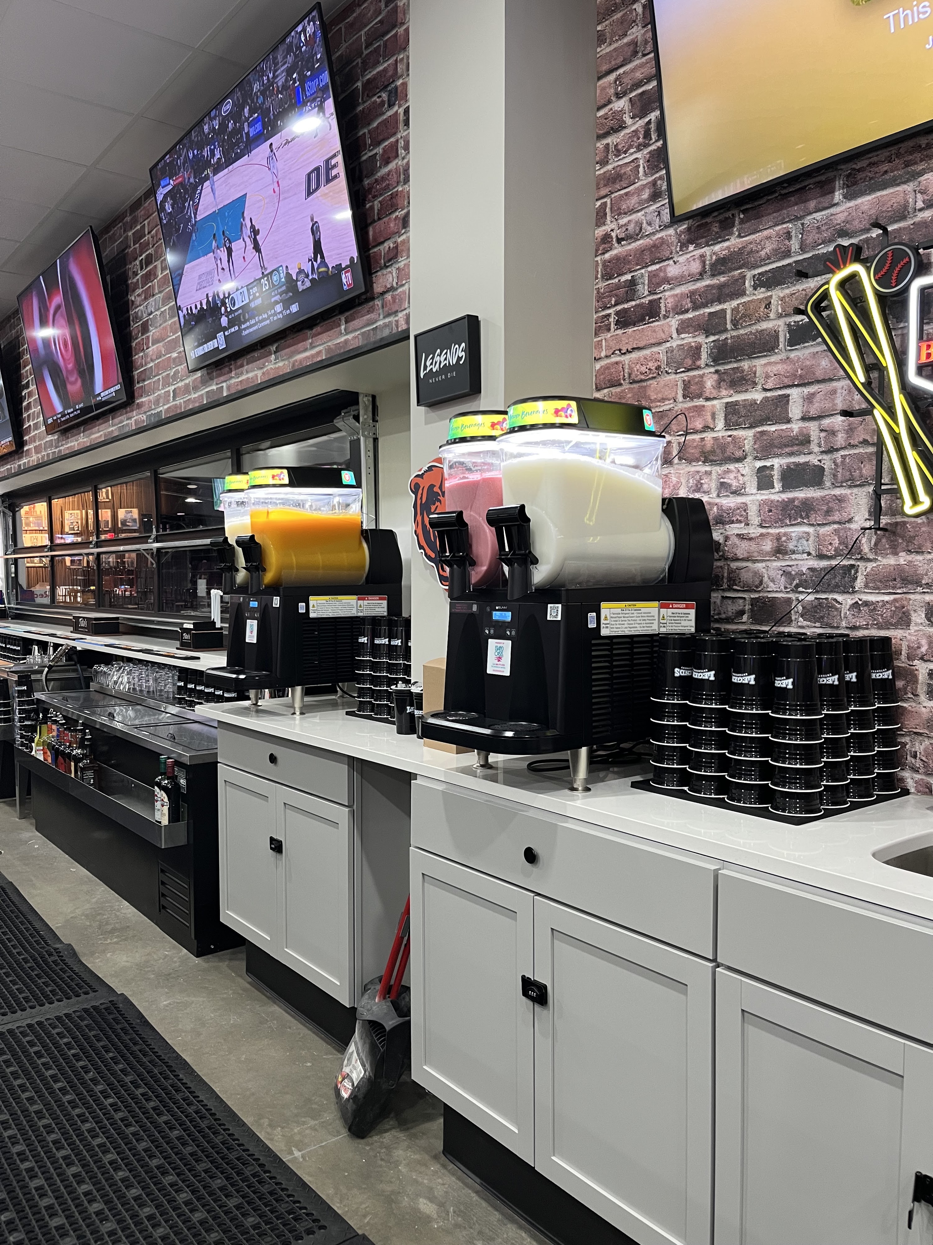 A drink station with slushy machines and stacked black cups against a red brick wall with televisions.