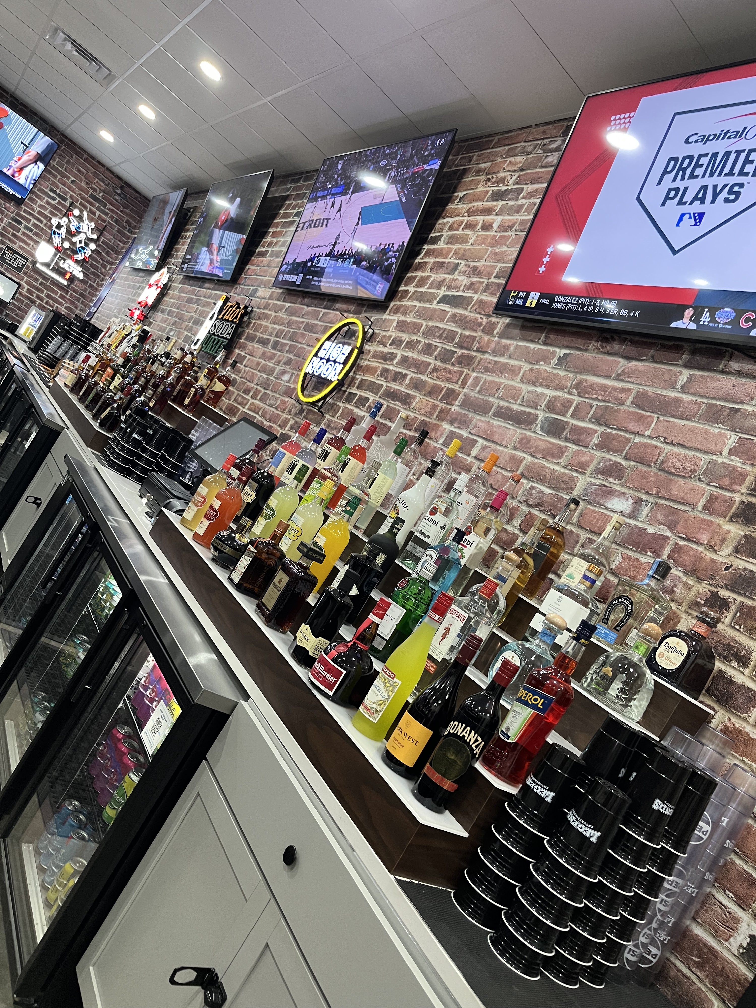 A well-stocked bar with numerous liquor bottles, beer coolers, and televisions mounted on a brick wall.