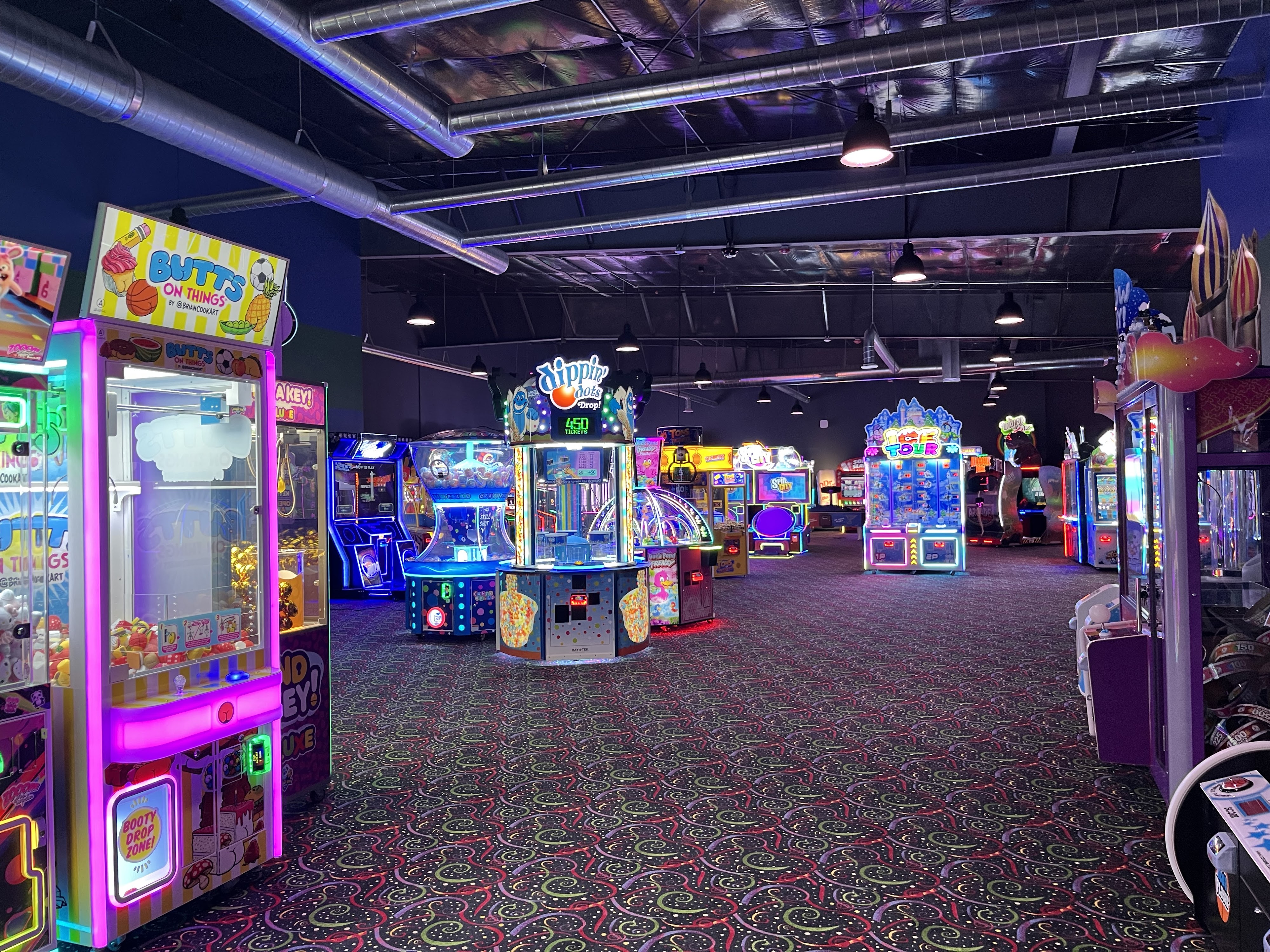 A brightly lit arcade room with various colorful claw machines and redemption games on a patterned carpet.