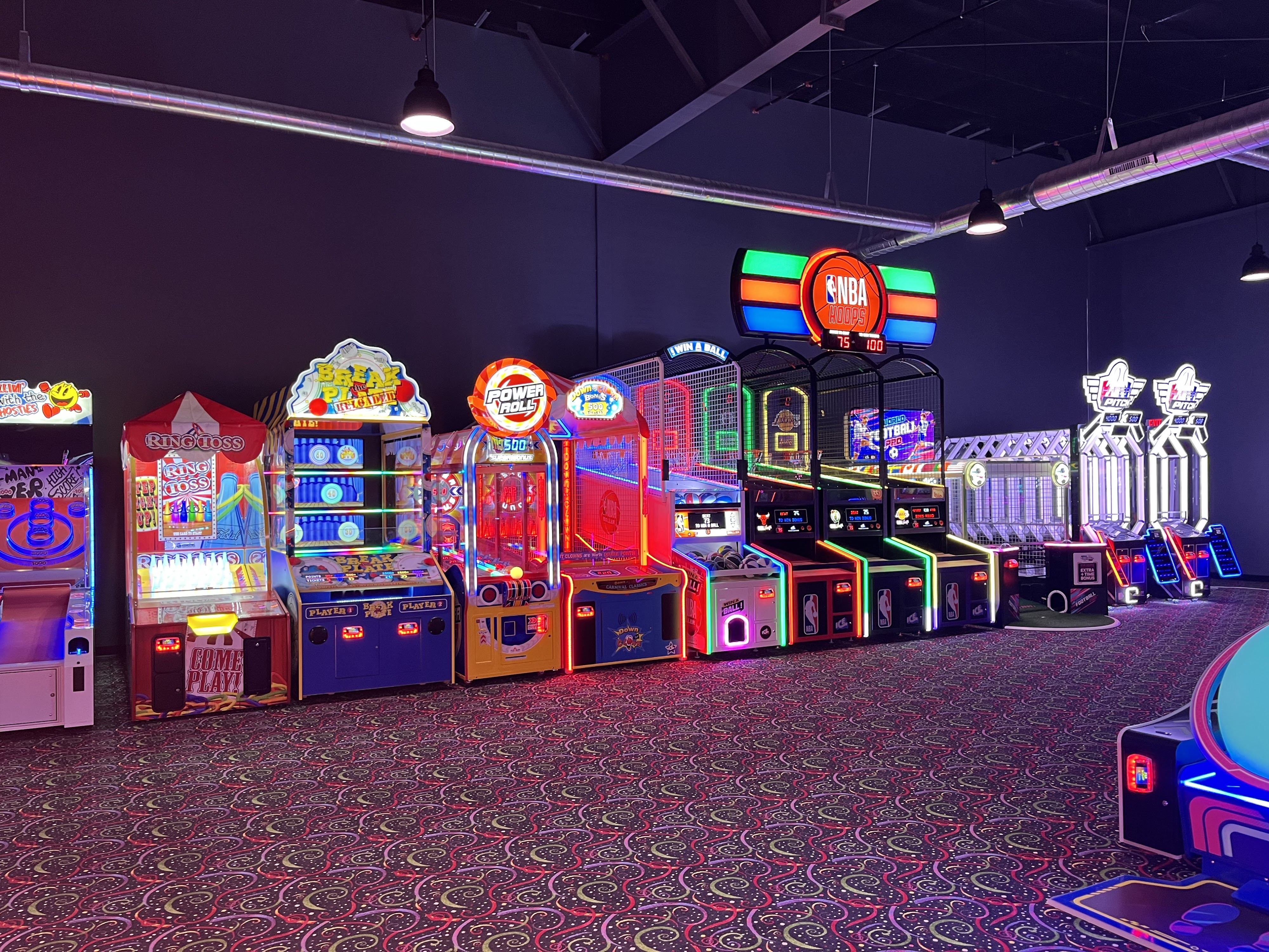 A row of colorful illuminated arcade games including basketball and ring toss in a dimly lit entertainment center.