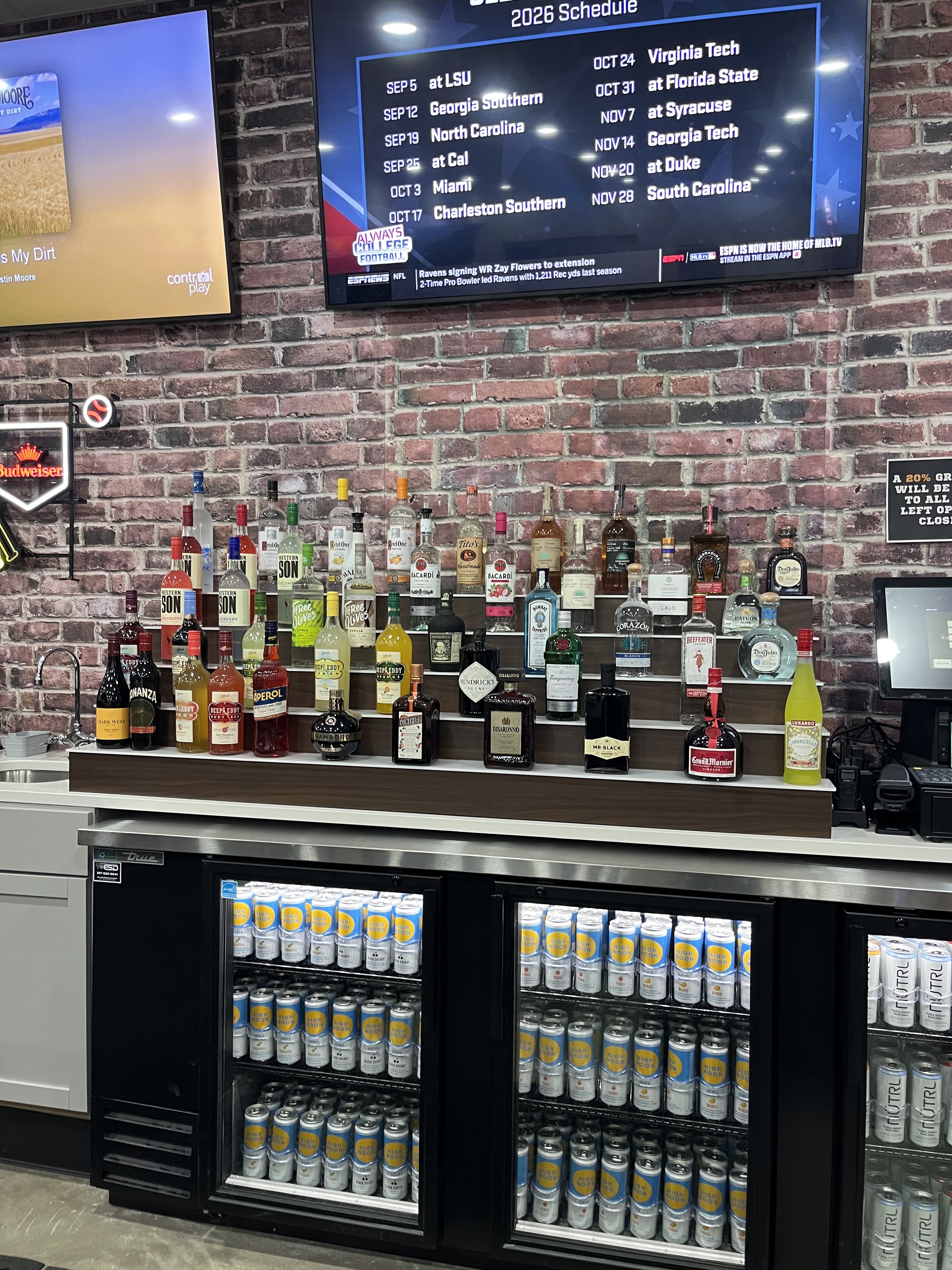 A well-stocked bar with three tiers of liquor bottles, coolers filled with canned seltzers, and a brick wall background.