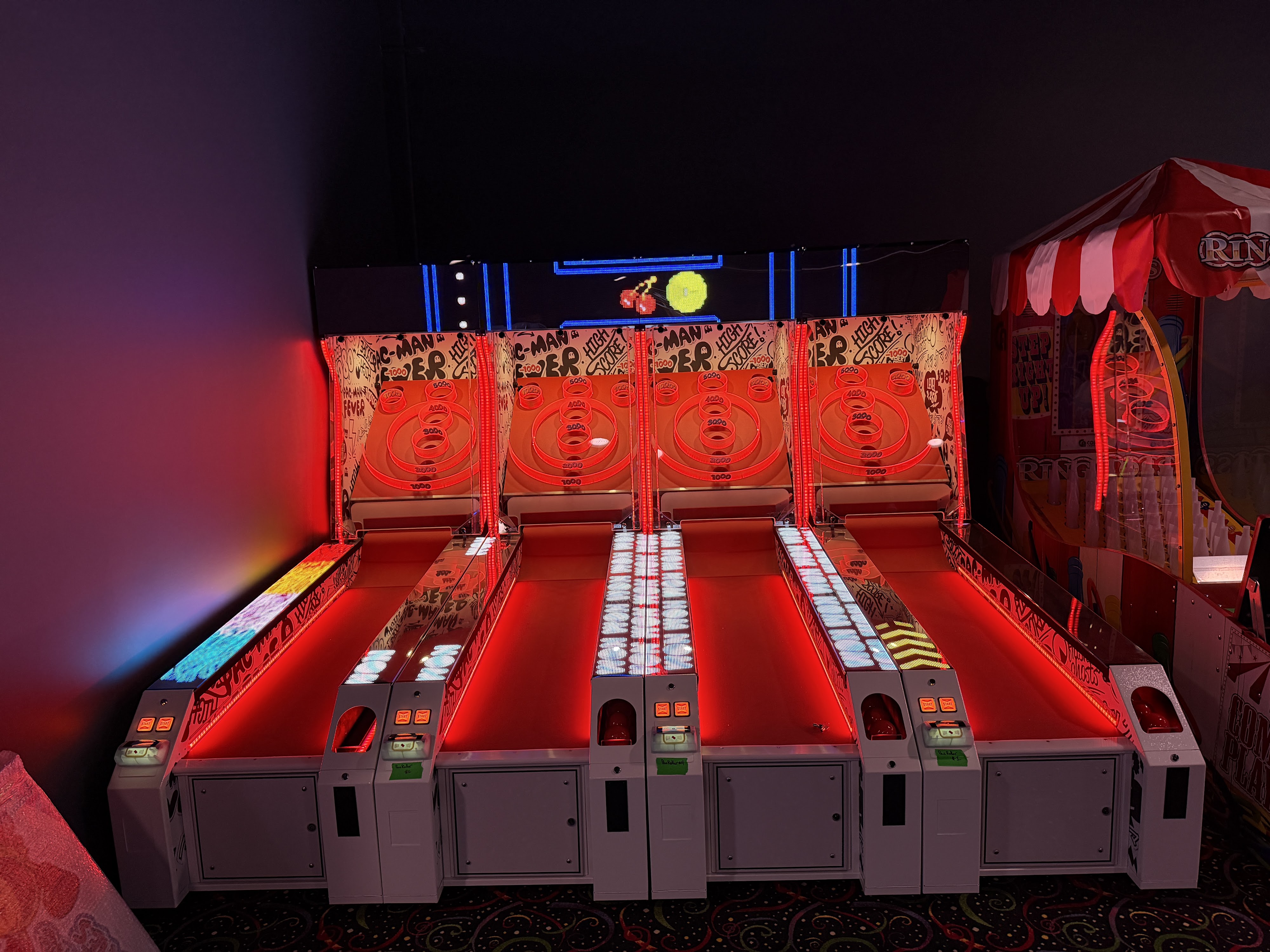 Four modern Skee-Ball machines with glowing red lights and Pac-Man graphics in a dimly lit arcade.