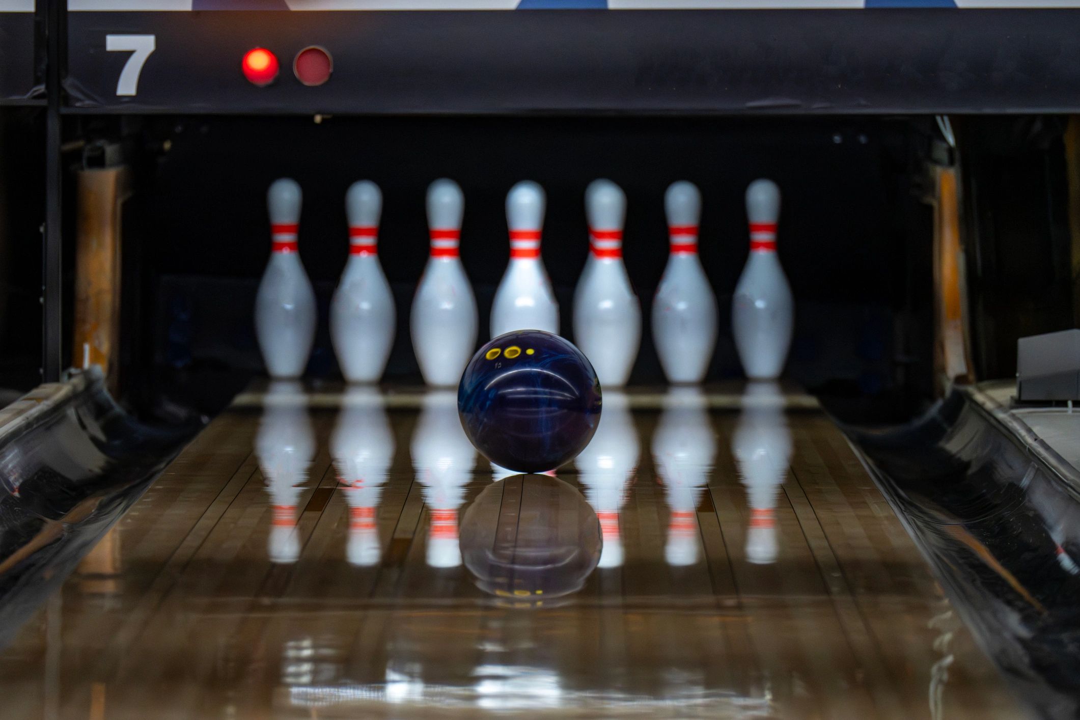 A blue bowling ball rolls down a shiny wooden lane toward white pins with red stripes on lane seven.