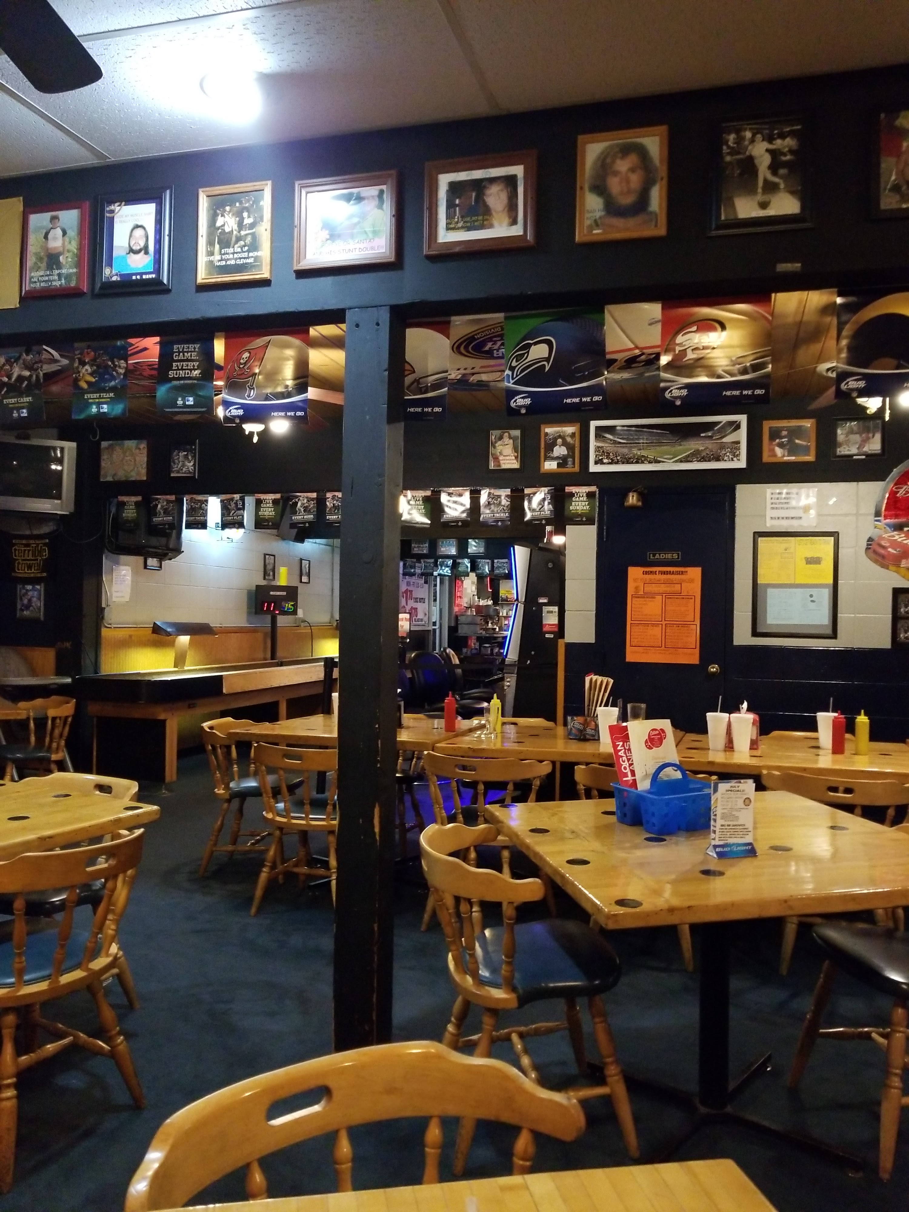Game-room dining area with wood tables and chairs, blue walls covered with framed photos and sports memorabilia, arcade machines, and serving counter.