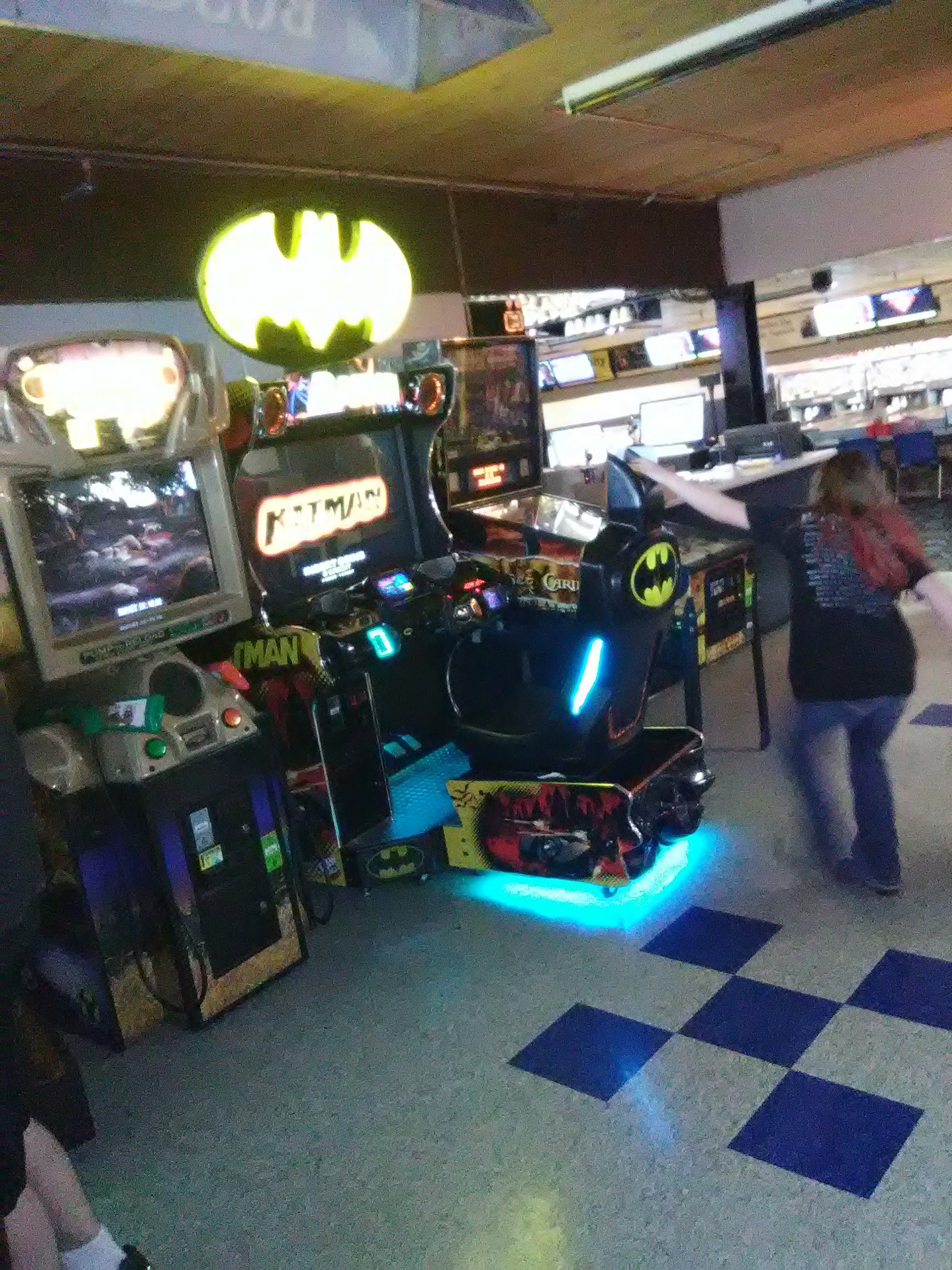 Batman-themed arcade game with cyan neon lighting and glowing bat sign, surrounded by other arcade cabinets in a bowling center.