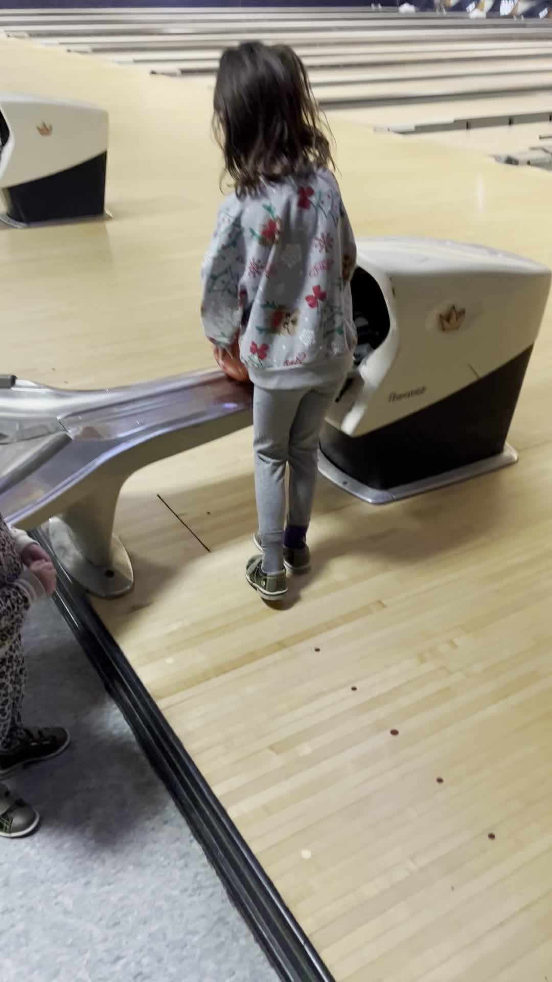 Child in floral jacket standing on bowling lane, ready to bowl.