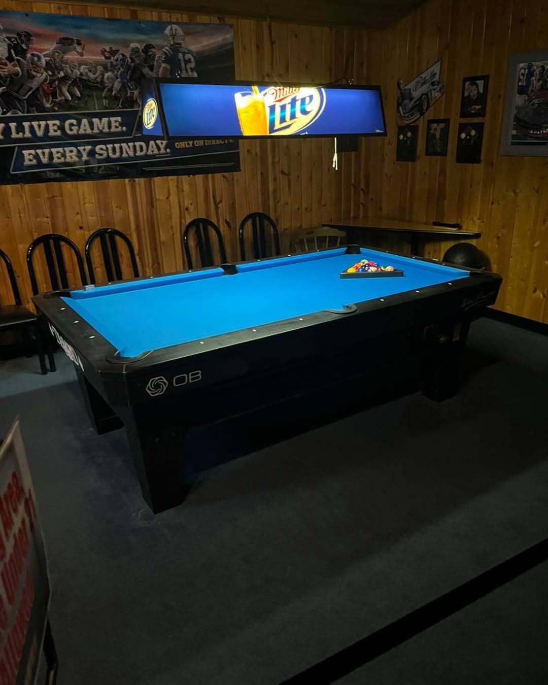 Blue pool table with racked balls in a wood-paneled room with Miller Lite sign and sports posters.