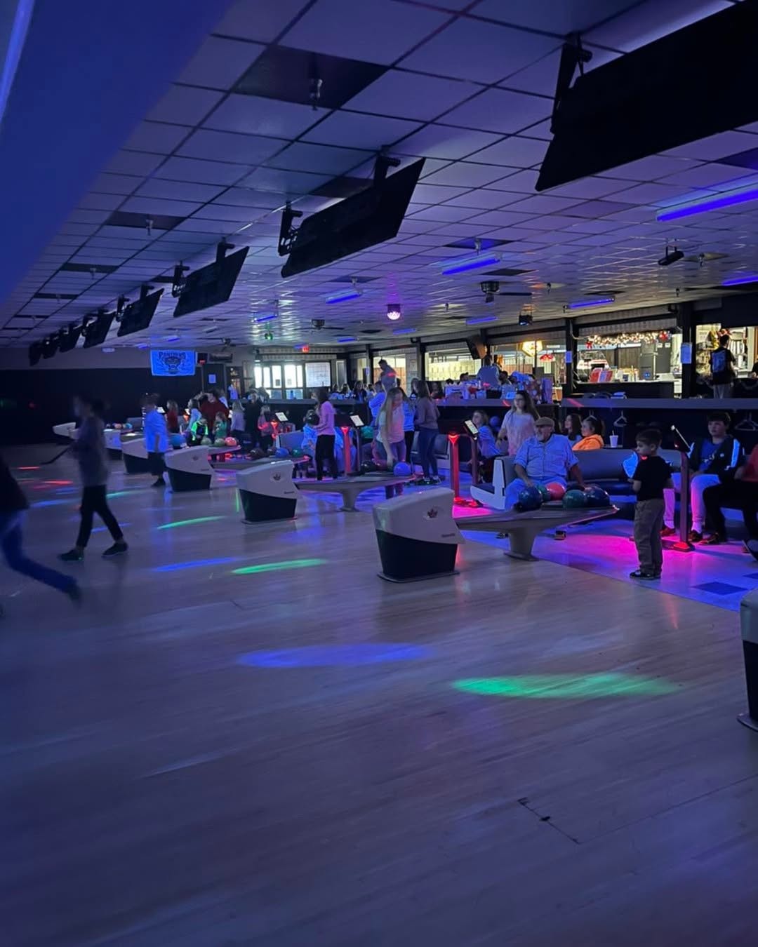 A bowling alley during cosmic bowling with neon lights reflecting on the lanes and people playing.