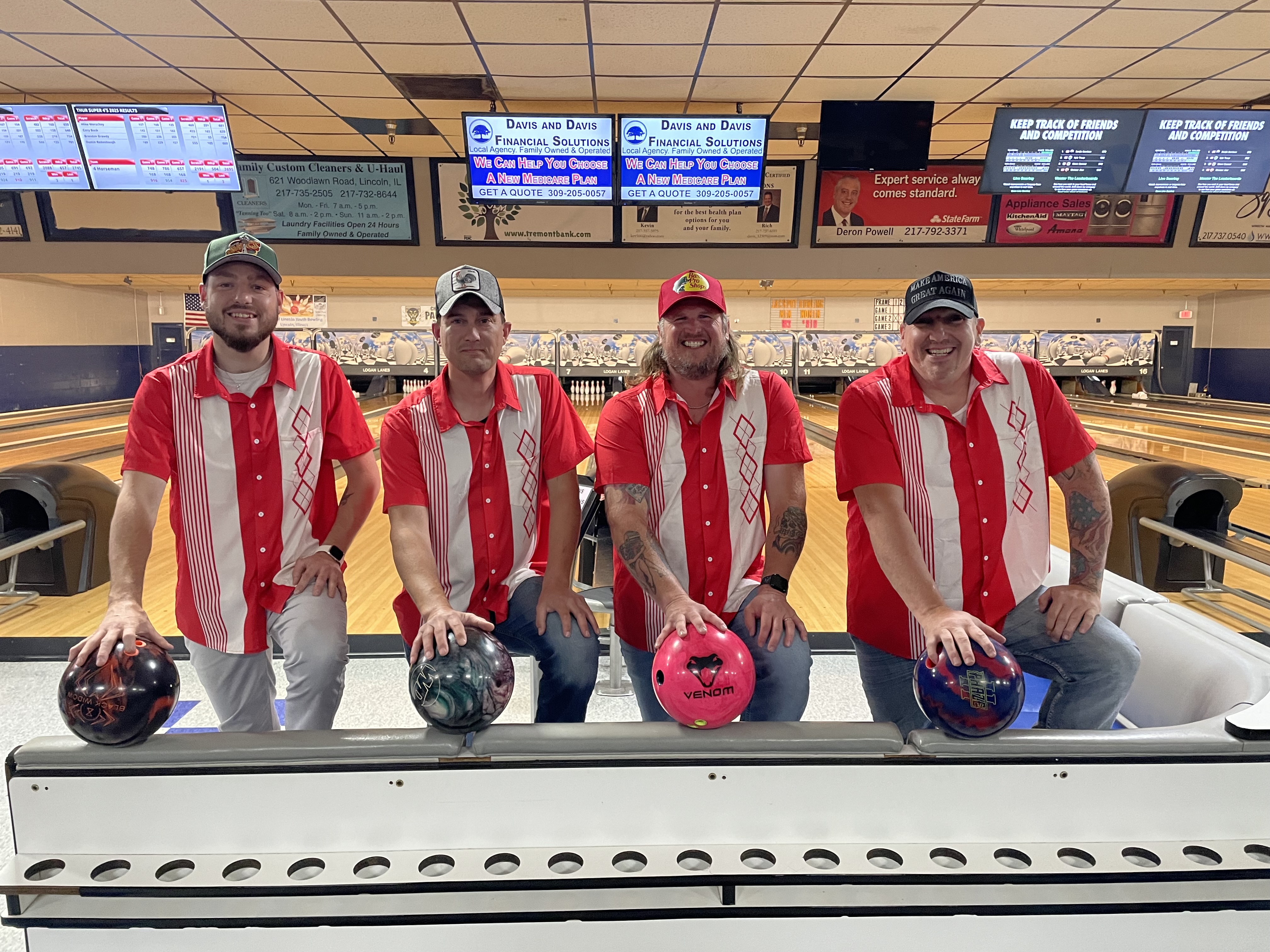 Four men in red and white bowling team shirts hold bowling balls at the score desk of a bowling center.