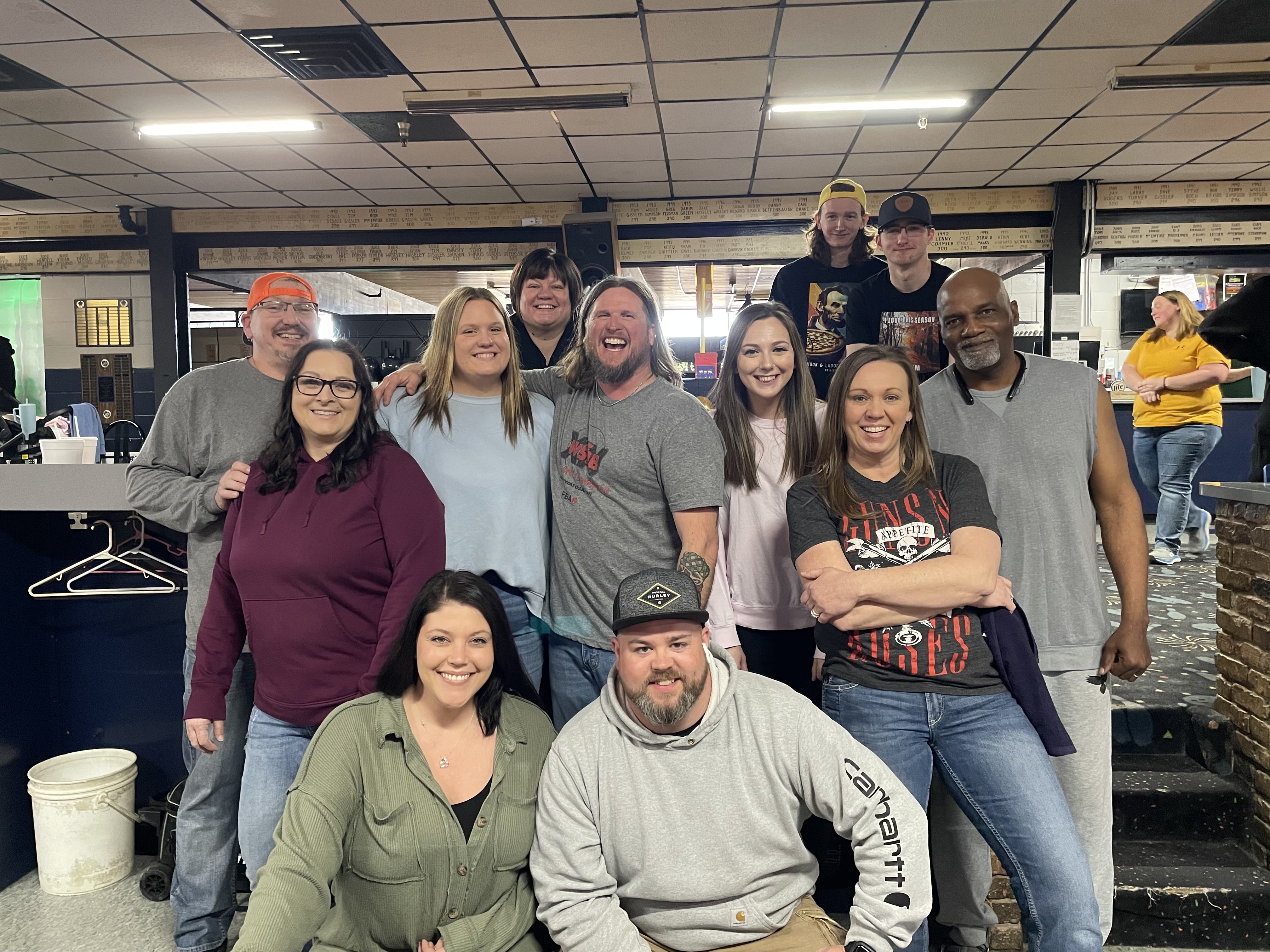 Group of 15 smiling adults posing together inside a bowling center.