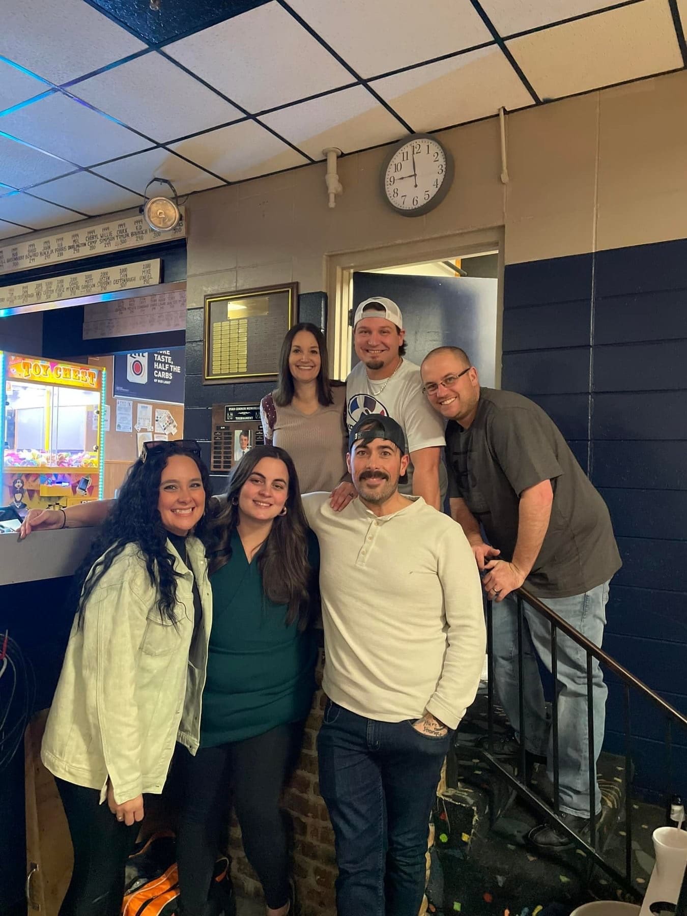 Six smiling adults posing together in a bowling center party room with arcade game and wall clock visible.
