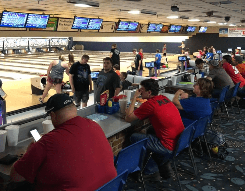 Busy bowling center with teams in red and blue shirts bowling and waiting at scoring tables during an organized event.