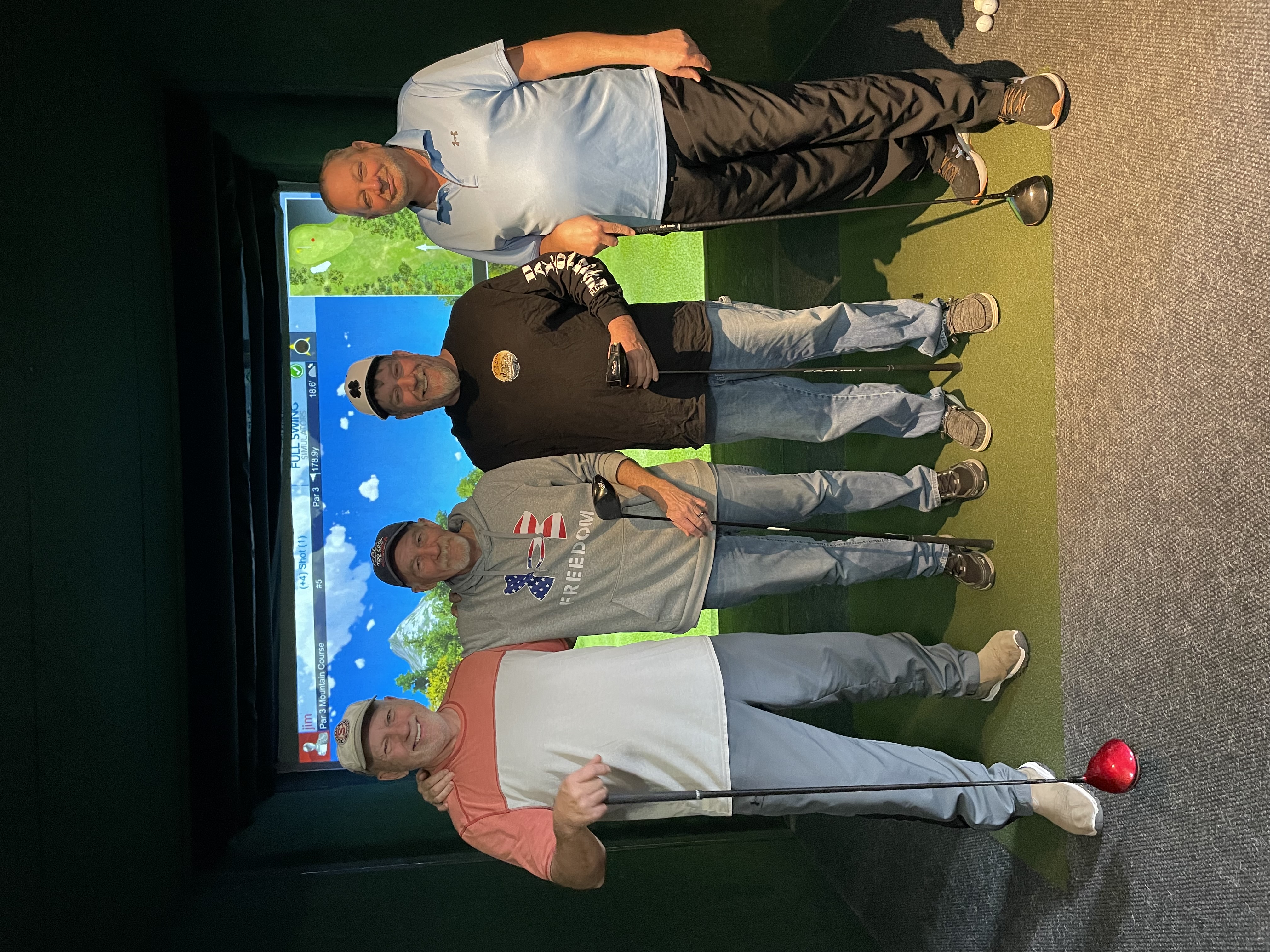 Four smiling men pose together in a golf simulator bay holding clubs in front of an active course display.