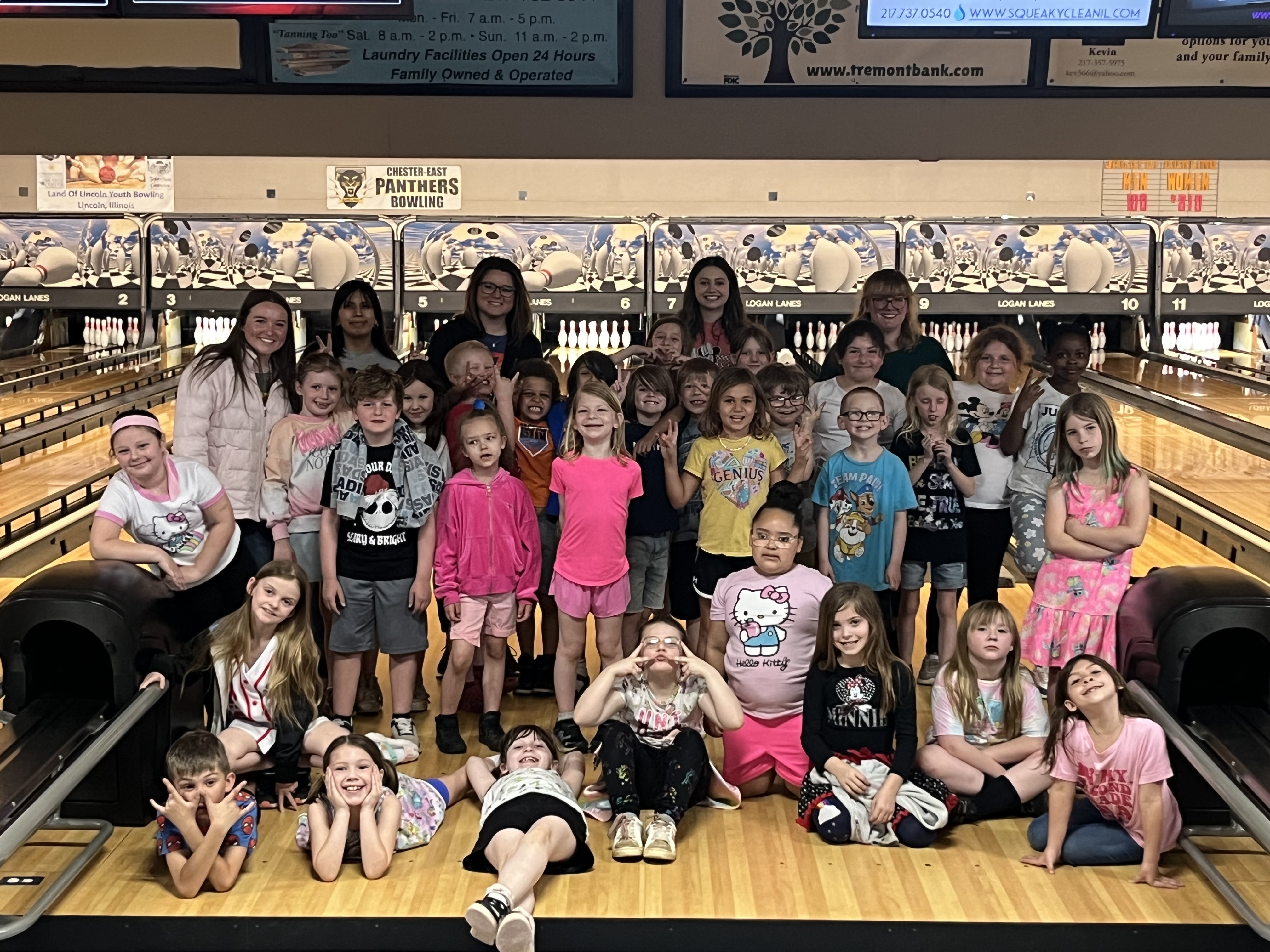 Large group of children and teens smiling at a bowling alley on lane flooring with pinsetters visible behind them.