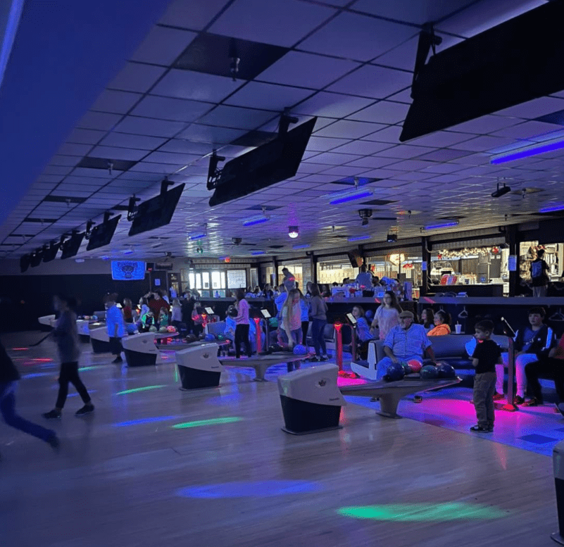 Children and adults bowl under vibrant blue and pink neon lights at a cosmic bowling alley.
