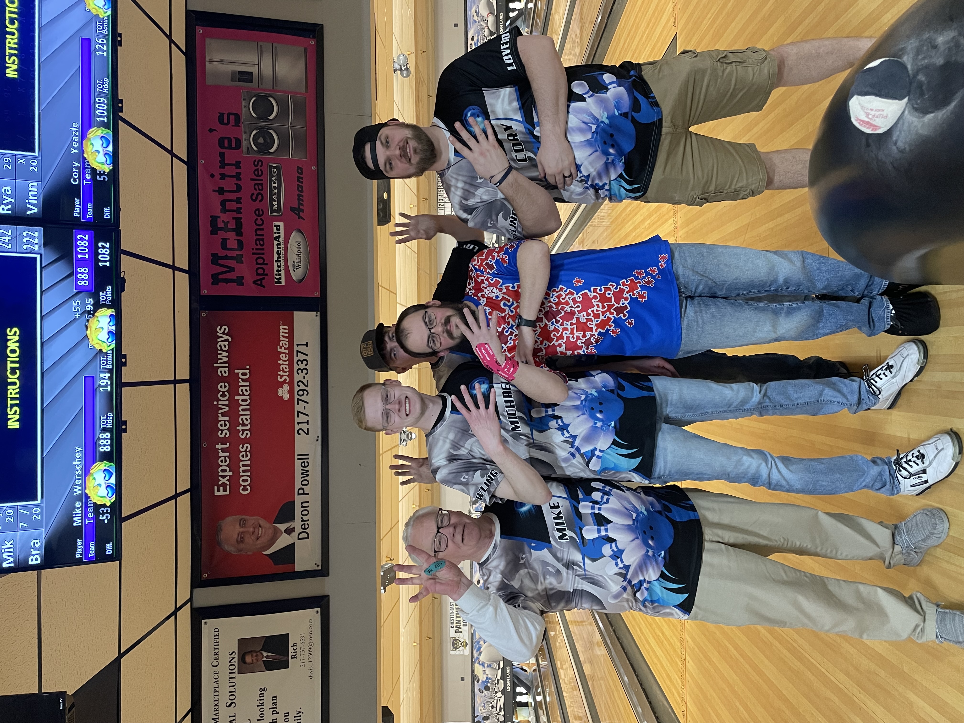 Five smiling bowlers in custom blue and black shirts pose together on a lit bowling lane with overhead scoreboards visible.
