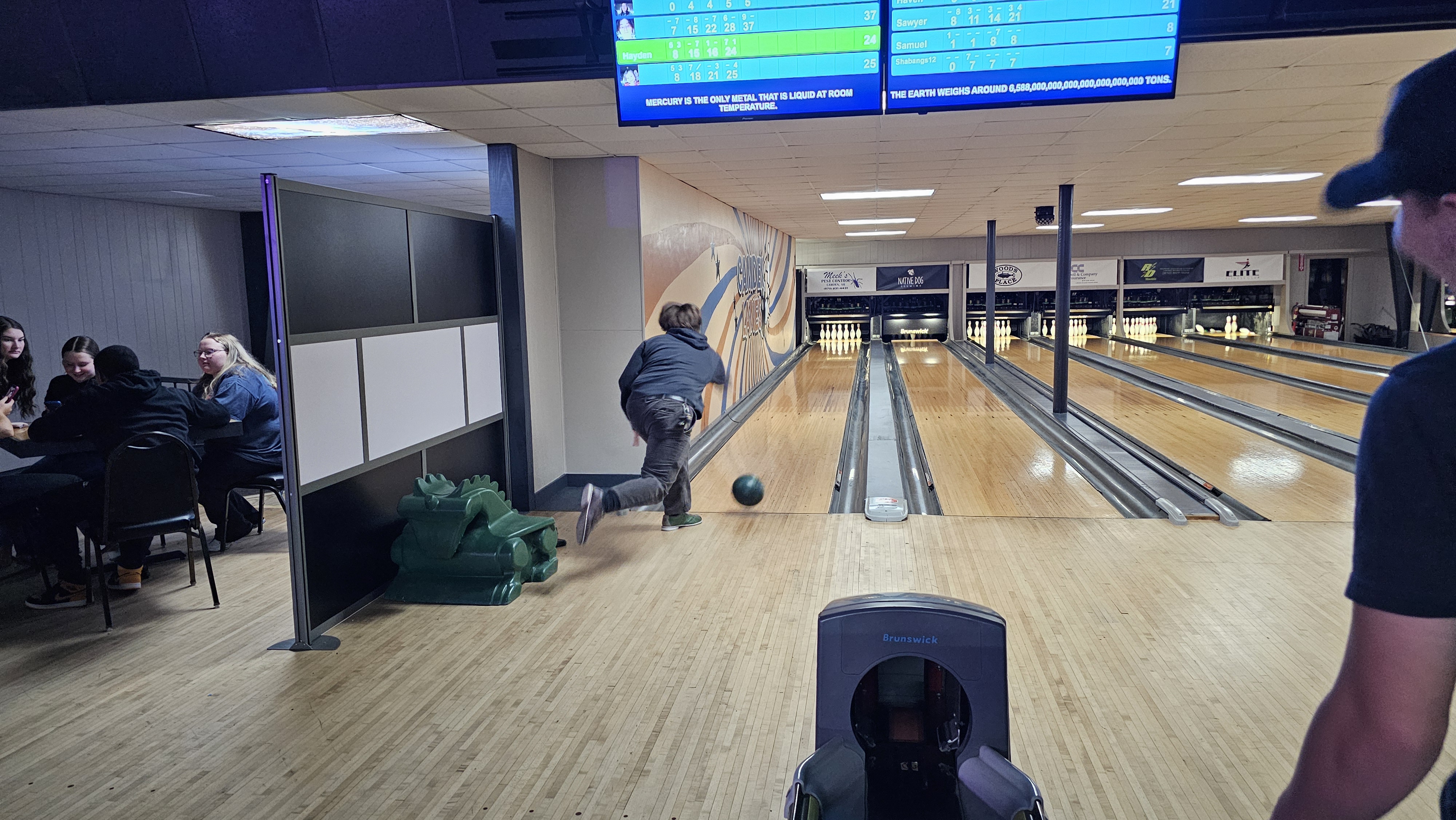 A person bowls a green bowling ball down a lane, with digital scoreboards above showing player scores and multiple lanes in the background.