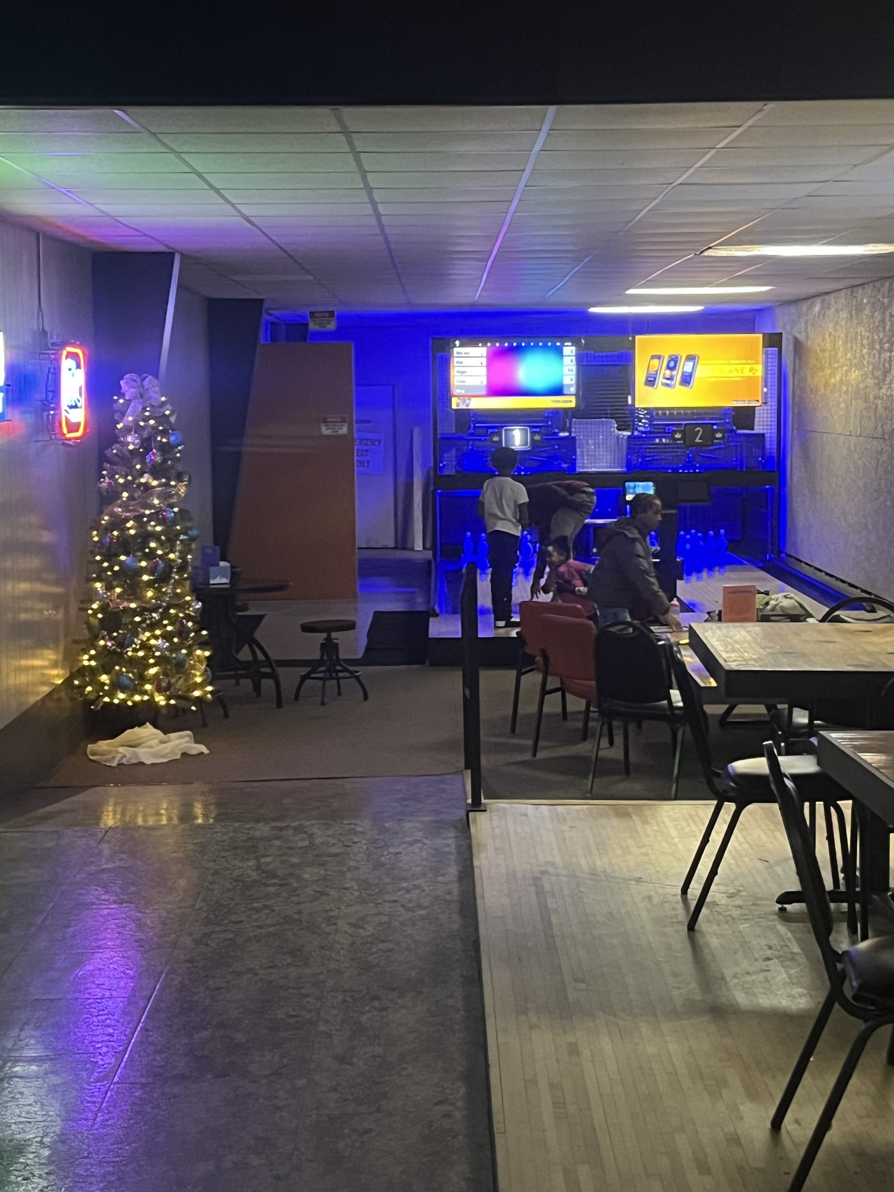 A bowling alley at night with blue and purple lighting, featuring a Christmas tree and people bowling.