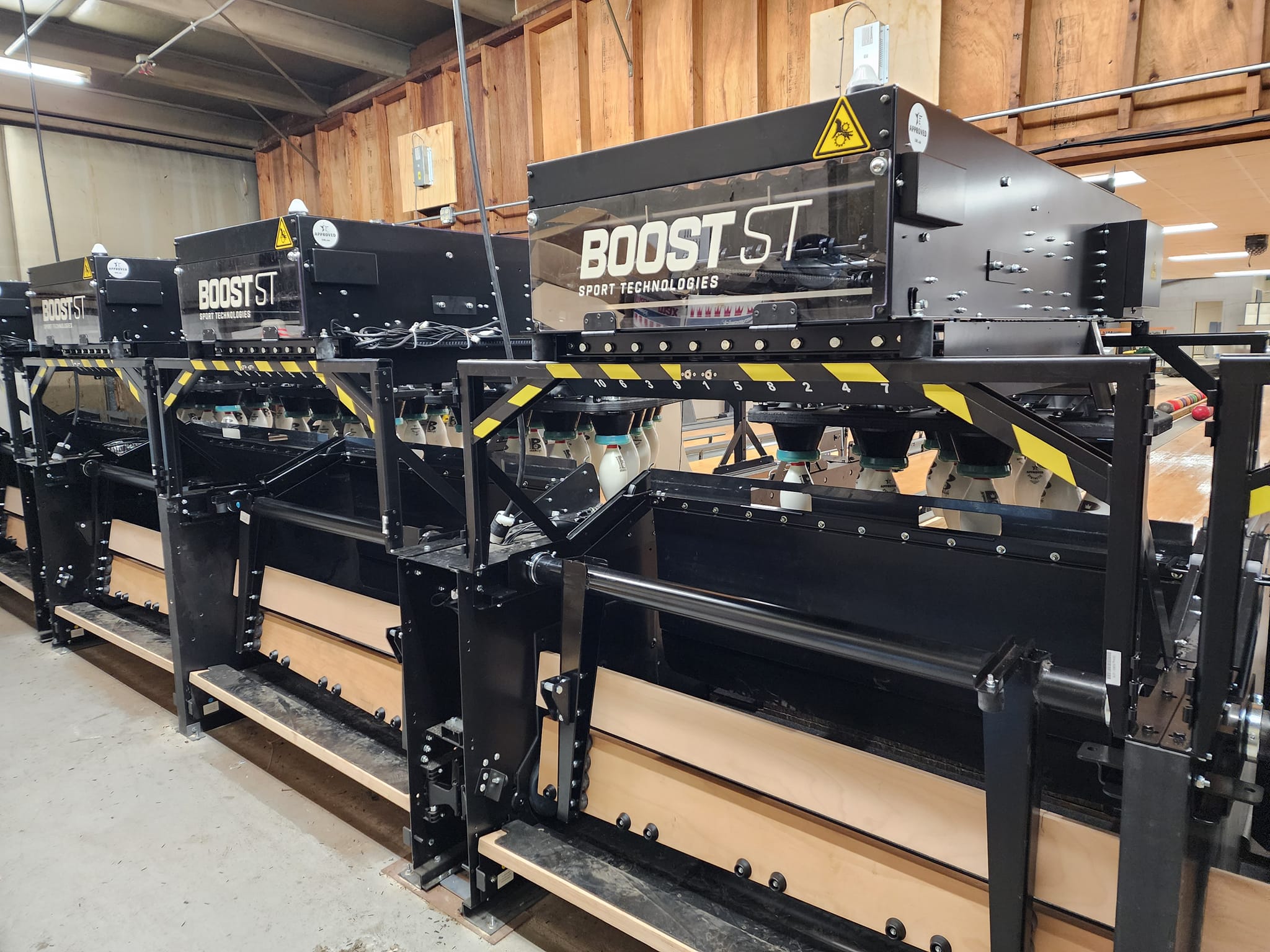 Close-up of several black bowling pinsetters with "BOOST ST Sport Technologies" branding, set against a wooden wall and industrial ceiling.