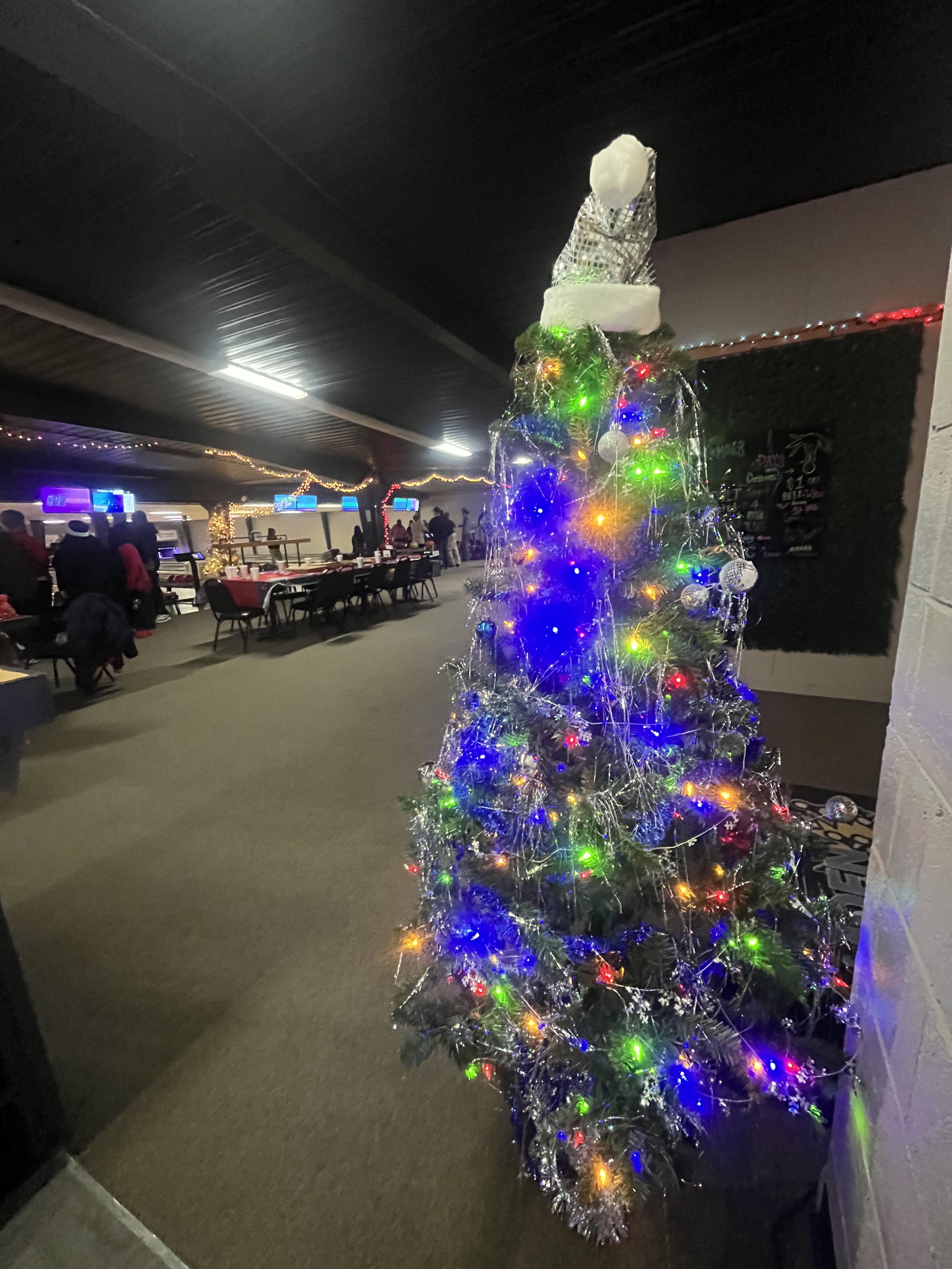 A festive Christmas tree with multicolored lights stands in the foreground of a dimly lit bowling alley with people and decorated tables.