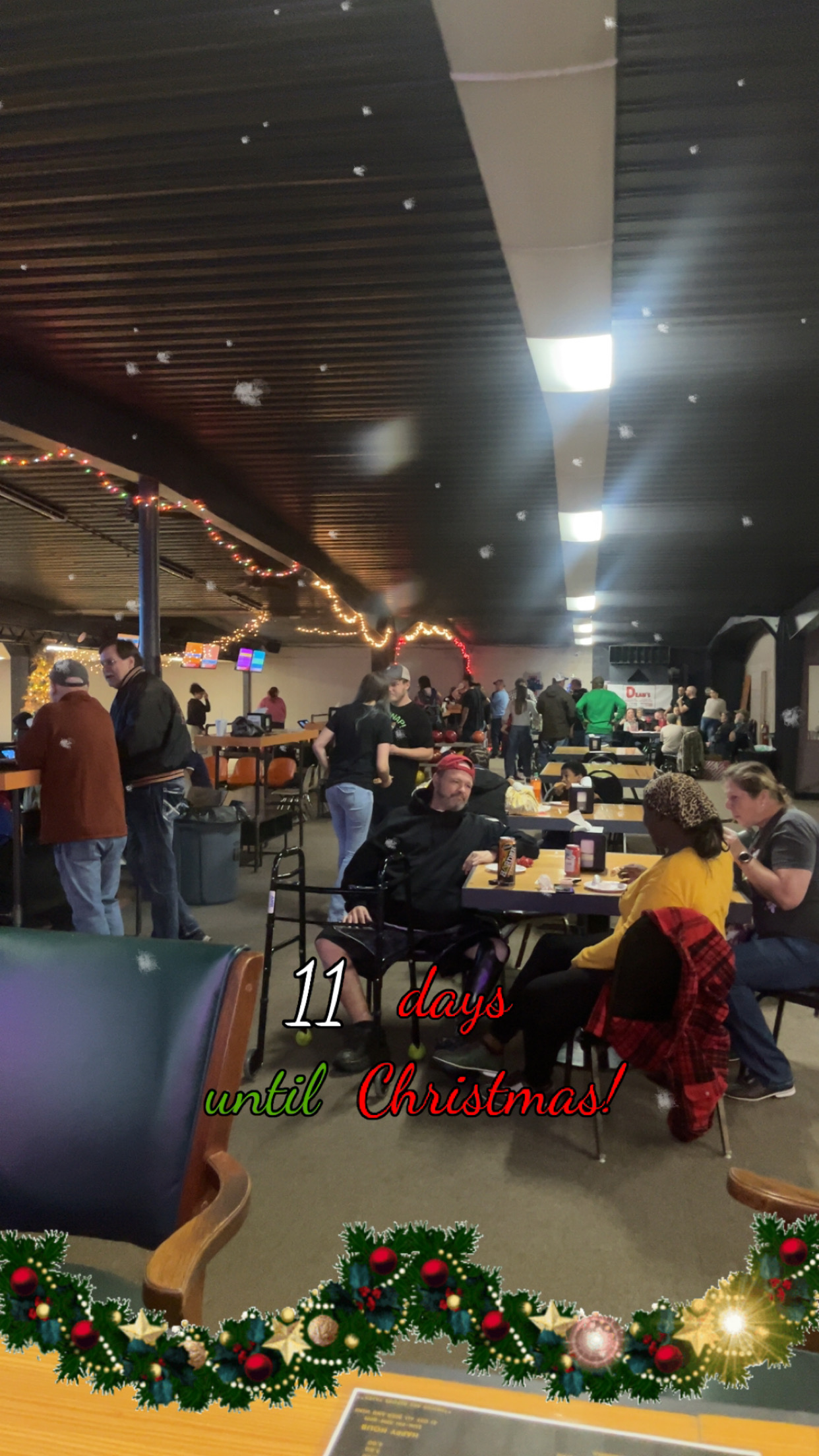 An indoor shot of a bowling alley with patrons sitting at tables and standing near lanes, decorated with Christmas lights and a garland with overlay text.