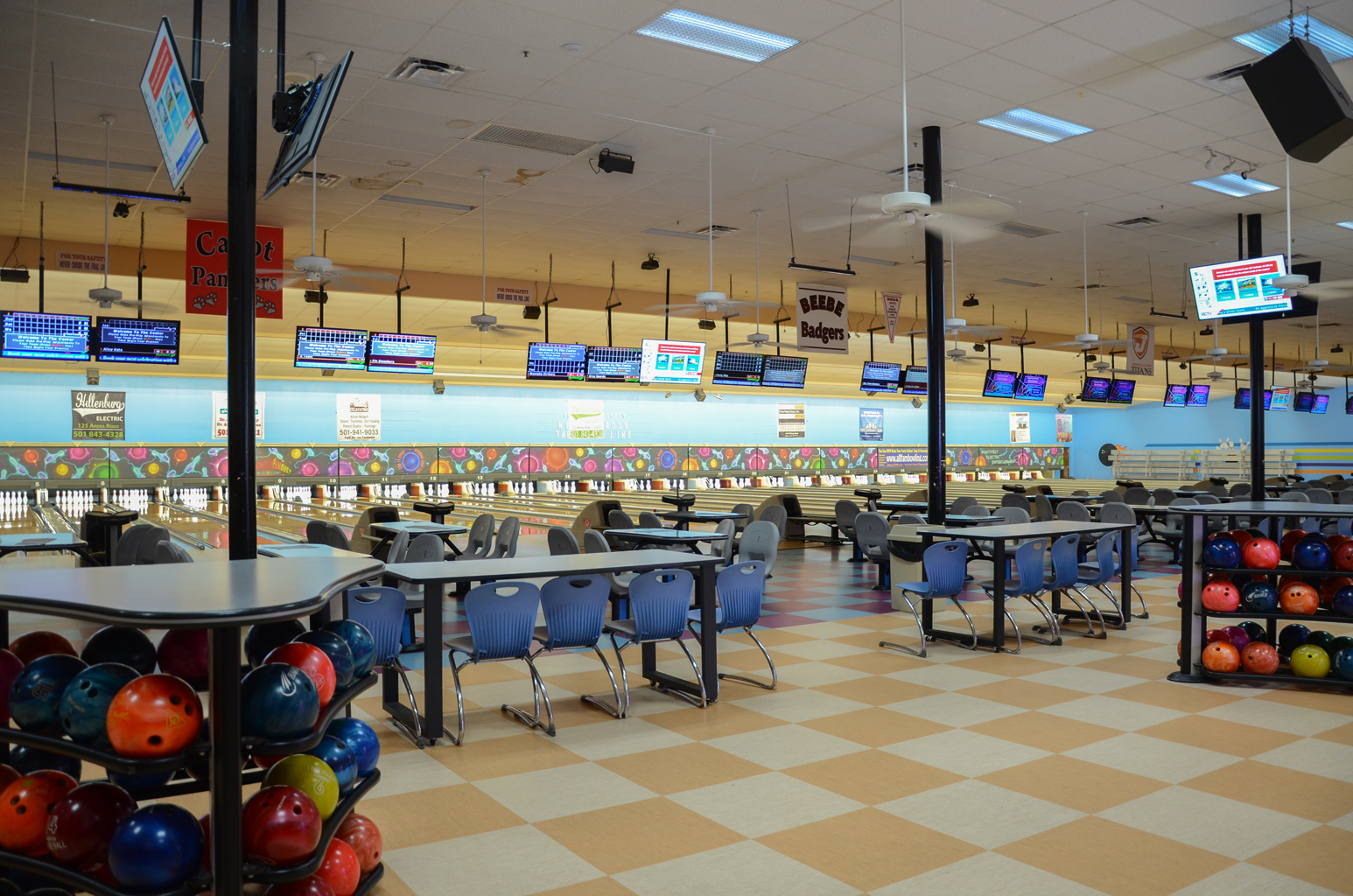 A wide interior view of a bowling alley with rows of lanes, blue seating areas, and racks of colorful bowling balls.
