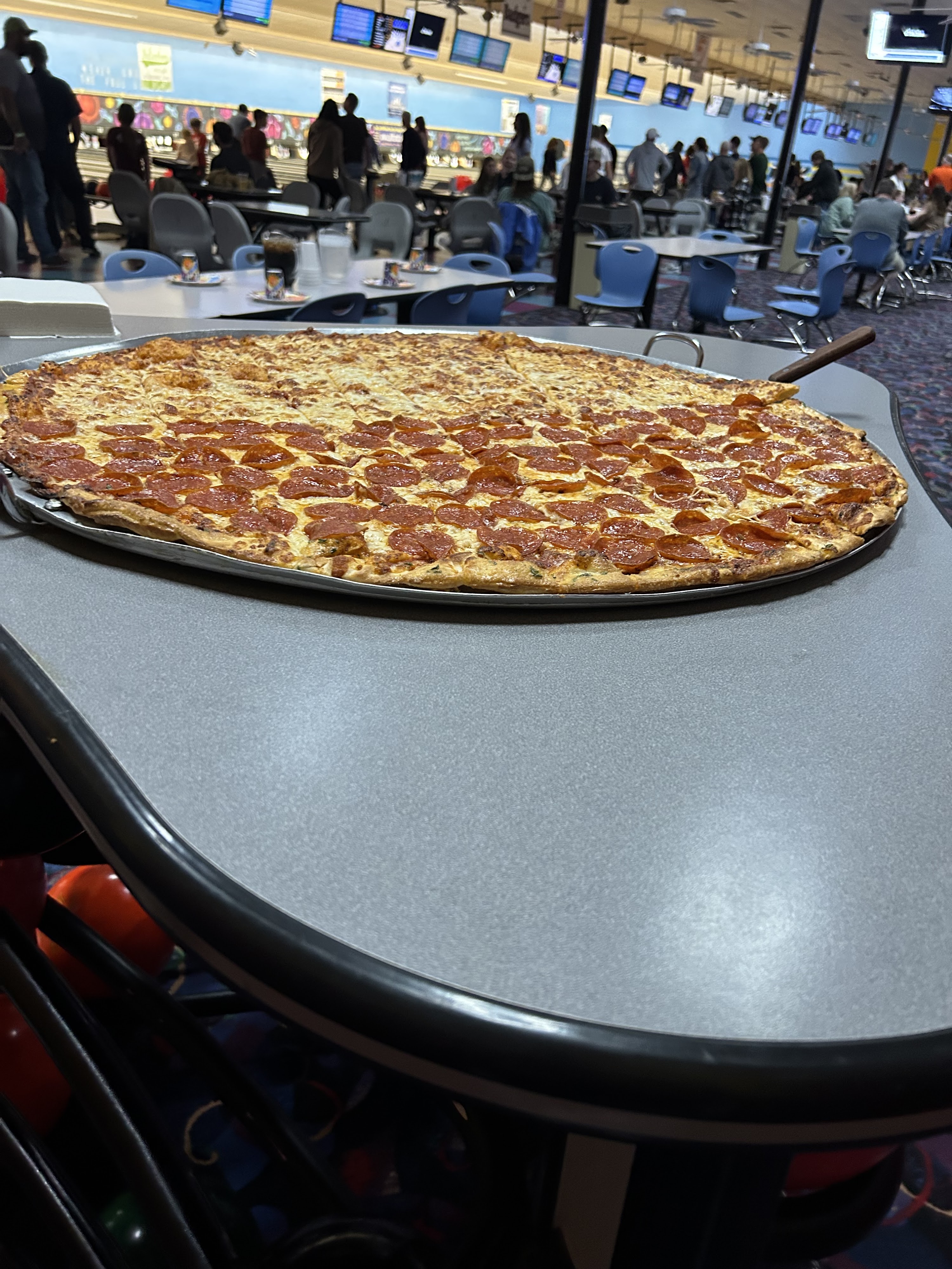 A large sheet pizza with pepperoni and cheese toppings on a table in a busy bowling alley.