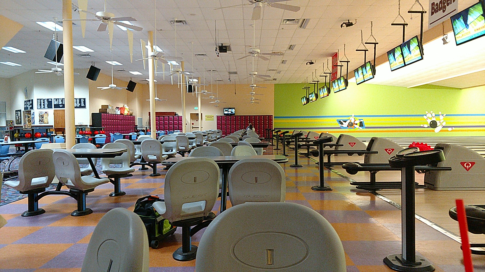 A brightly lit bowling alley interior with grey seats, checkered floors, and several lanes stretching into the distance.