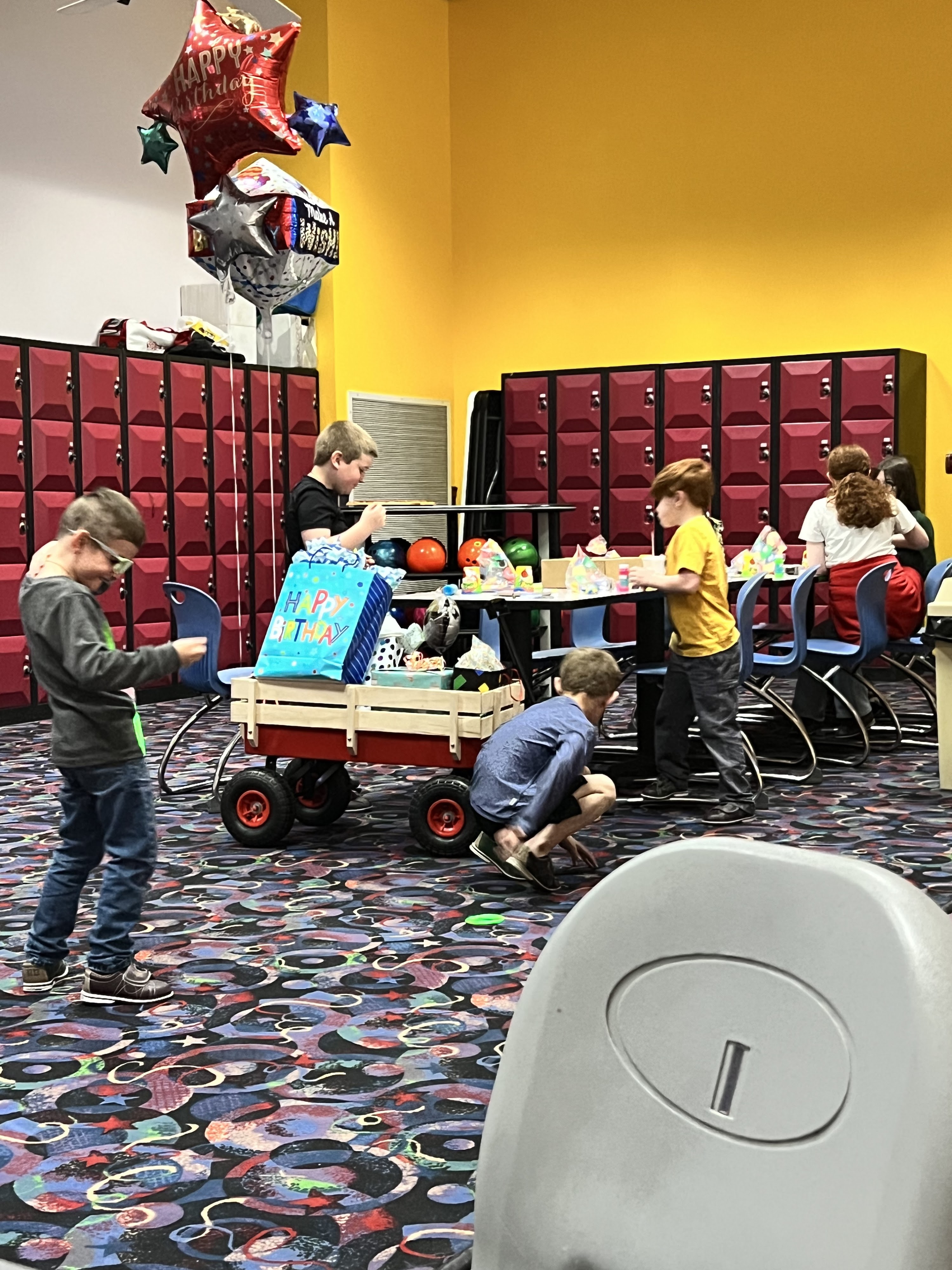 Children celebrate at a birthday party in a bowling center with a gift wagon, balloons, and colorful carpet.