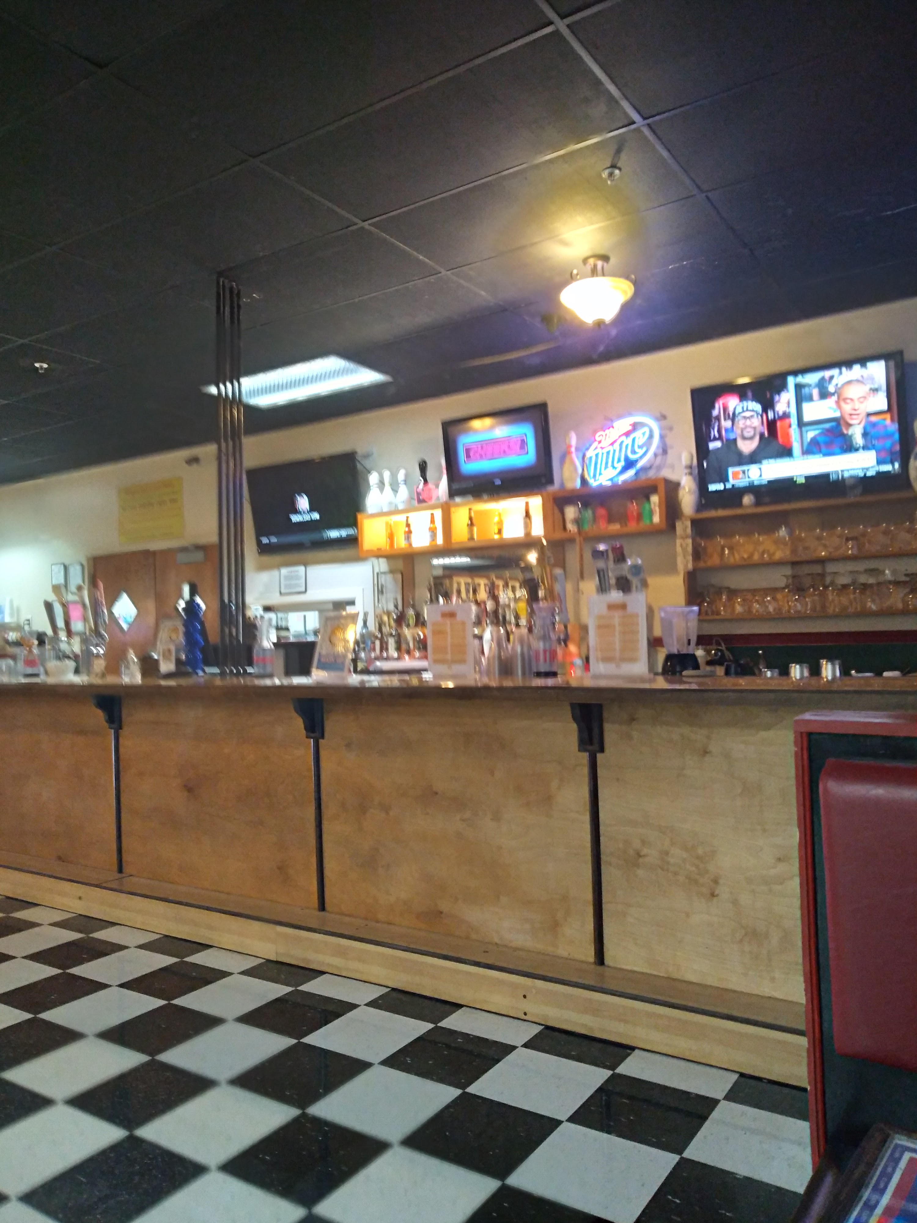 A wooden bar counter with liquor bottles, neon signs, and three televisions above a checkered floor.