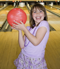 A young girl smiling while holding a red bowling ball on a bowling lane.