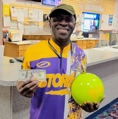 A man in a purple and yellow bowling jersey holds a neon yellow bowling ball and a hundred-dollar bill.