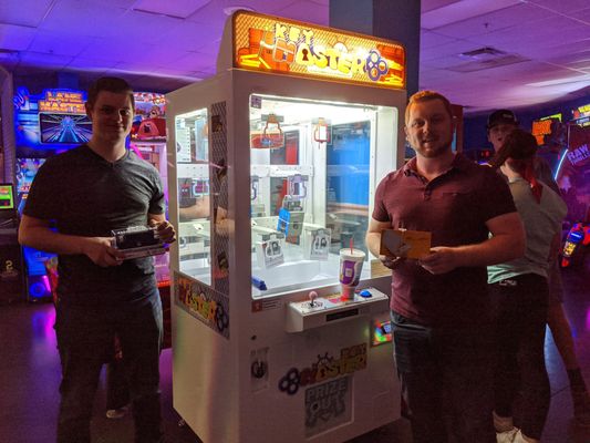 Two men pose with small boxed prizes in front of a glowing Key Master arcade game.