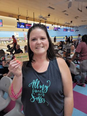 A smiling woman holds a rewards card inside a bowling alley with lanes and other bowlers in the background.